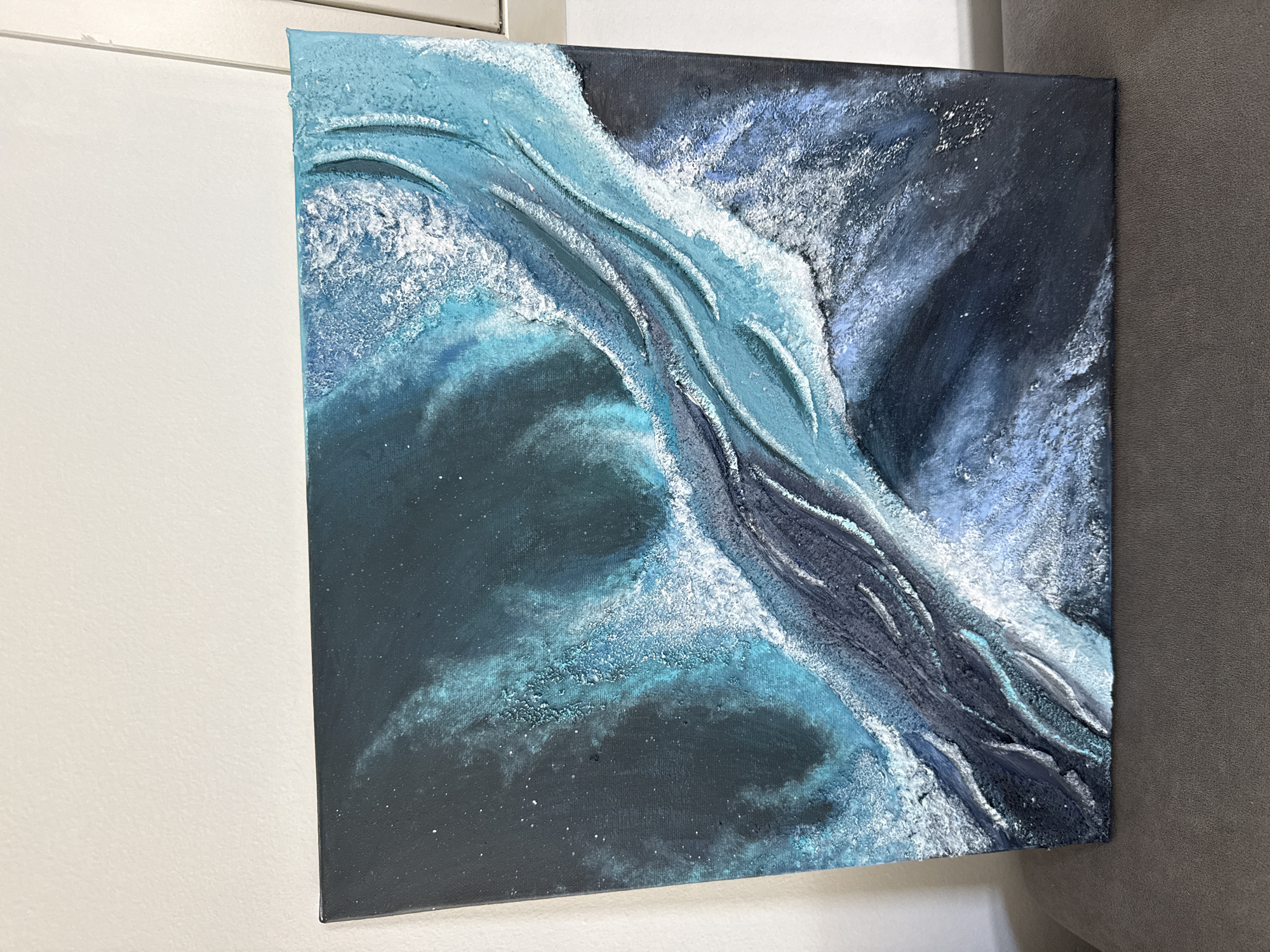 The image shows a textured abstract painting with a wave-like motif in shades of blue and white. The painting is placed on a dark grey surface, possibly a sofa or chair, in front of a white wall. The lighting suggests an indoor setting, with natural light coming from the right. There are no people or identifiable landmarks from Limassol, Cyprus present in the image.