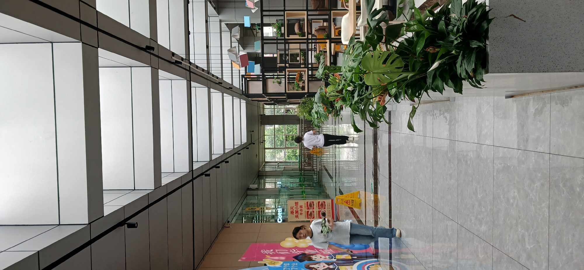 The image depicts an indoor walkway, likely within a commercial or public building in the Chongqing High-tech Zone, China. The walkway features a tiled floor and a modern ceiling with a series of translucent panels that allow natural light to filter through, supplemented by track lighting.
On the left side of the frame, a young woman wearing a white t-shirt and blue jeans walks towards the viewer, holding a bundle of clothes. Adjacent to her is a colorful mural or advertisement. Further down the walkway, another person, also wearing a white t-shirt and dark pants, walks away from the camera. A yellow "wet floor" caution sign is visible in the middle ground.
The right side of the walkway is lined with a large planter filled with lush green foliage, including large-leafed plants and smaller leafy plants with white flowers. Behind the plants, a black metal shelving unit is displayed, containing various decorative items, plants, and what appear to be seating areas. Large windows are visible in the background, offering a view of green trees and an outdoor area. The overall environment suggests a clean, well-maintained, and contemporary space.