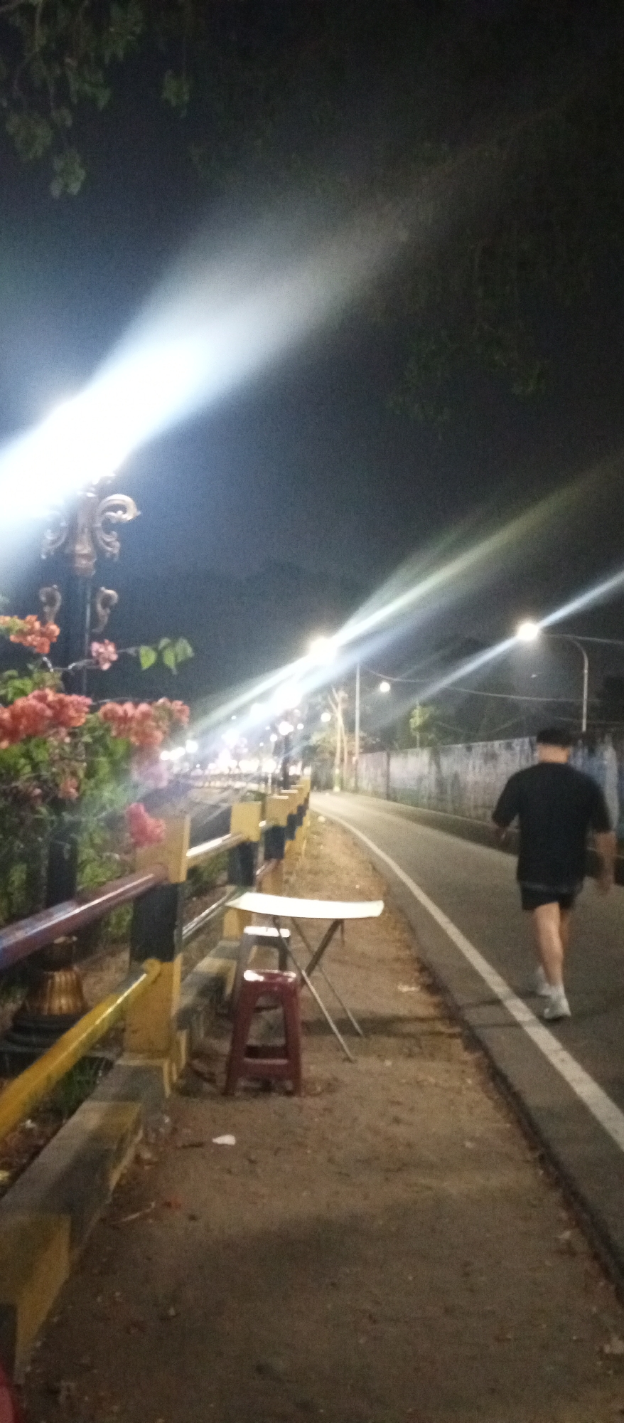 An outdoor scene at night in Madiun, Indonesia features a man walking on a paved path beside a road. He is wearing a black t-shirt, black shorts, and white sneakers. To his left, a yellow and black railing separates the path from a sidewalk lined with red and pink flowers. A red stool and a small white folding table are set up on the sidewalk. Streetlights cast bright beams across the scene, illuminating the road and trees in the background. The road appears to be a two-lane road with white lines marking the lanes. A concrete wall runs along the far side of the road. The sky is dark, suggesting it is nighttime.