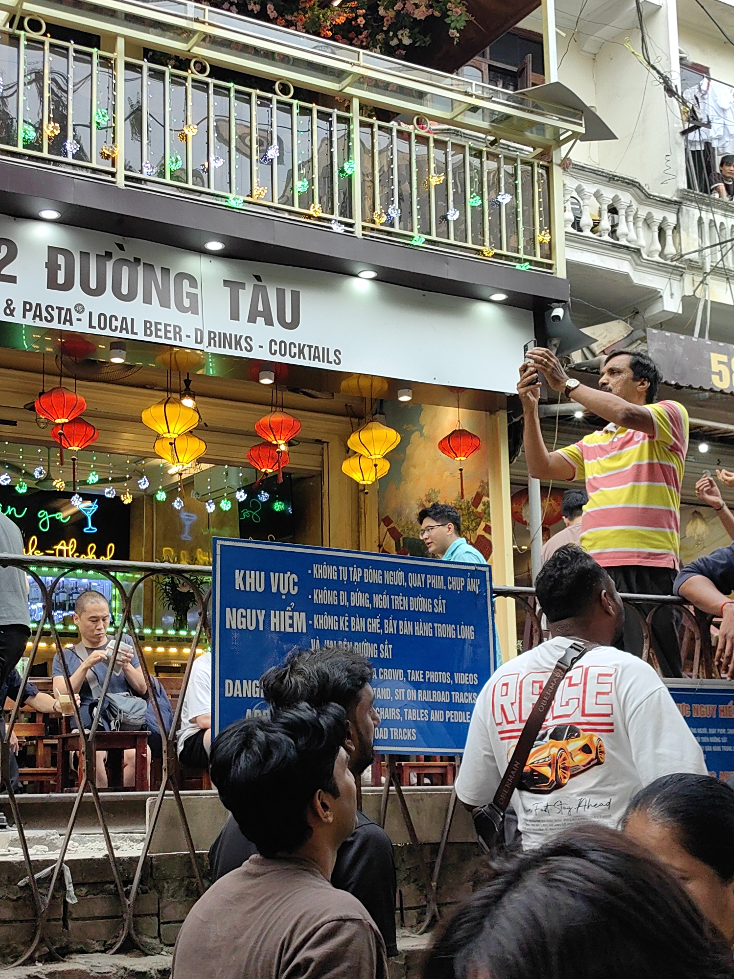 The image shows a crowded street scene in Hà Nội, Vietnam, in front of a restaurant named "2 Đường Tàu," which translates to "2 Train Street," offering local beer, drinks, and cocktails, and serving pasta. The restaurant has hanging red and yellow lanterns and decorative lights. A sign in Vietnamese and English warns about the dangers of gathering, filming, taking pictures, walking, standing, or sitting on the railroad tracks. Several people are present, including a man in a striped shirt taking a photo with his phone, a person in a "Race" t-shirt, and others seated at tables inside the restaurant. A man sits alone on a stool inside the restaurant looking at his phone. The overall scene appears to be a busy tourist attraction.