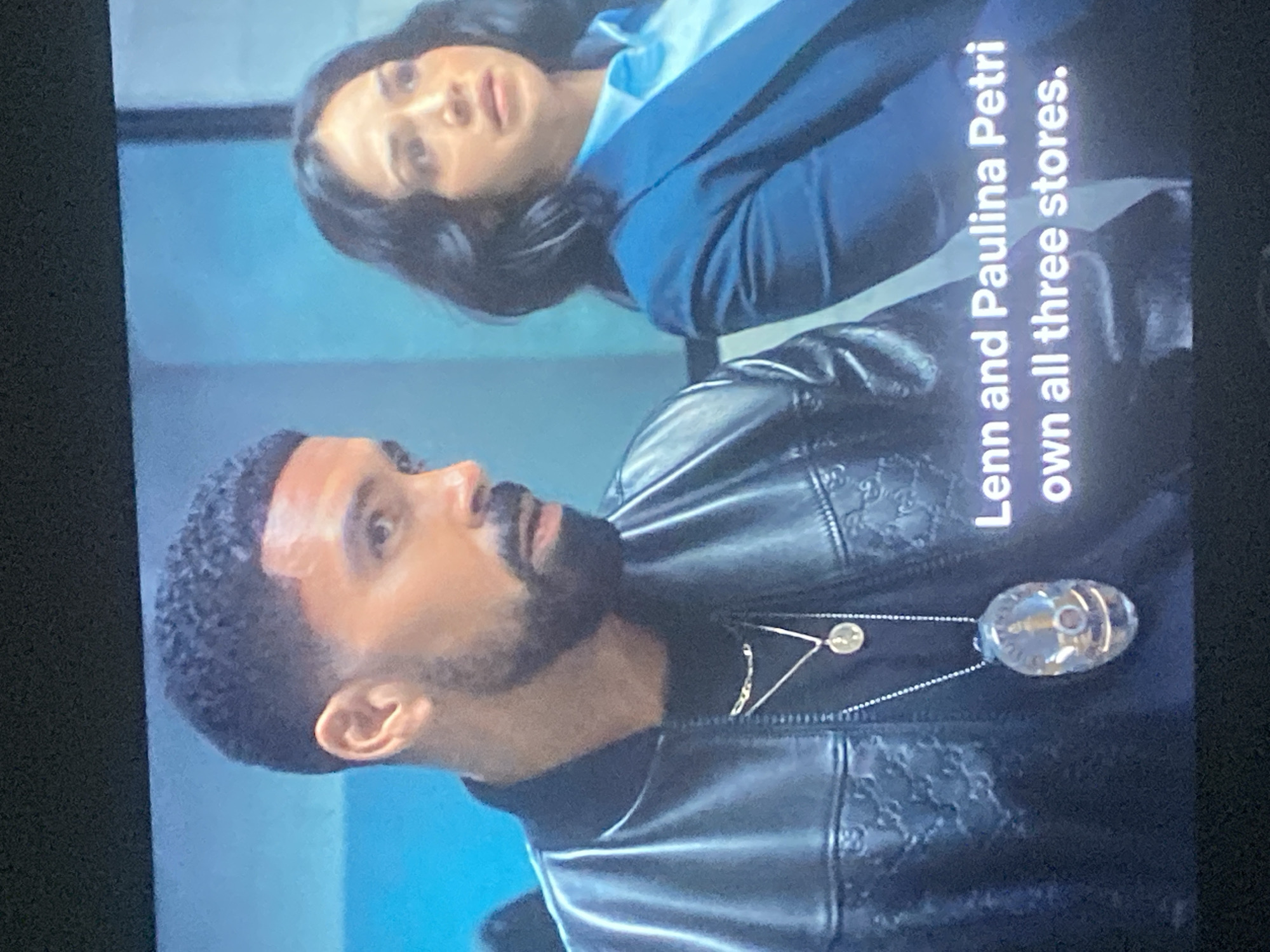 The image, originally rotated, displays two individuals—a man in the foreground and a woman behind him—in what appears to be an indoor setting with a plain light blue background.
The man, positioned on the left side of the frame, has dark, short, curly hair and a neatly trimmed beard. He is wearing a black, possibly leather, bomber-style jacket with a subtle, embossed pattern, resembling interlocking motifs (e.g., Gs) across the fabric. Underneath, he wears a dark shirt. He is adorned with two necklaces: a shorter one with a small circular gold-colored pendant, and a longer one featuring a larger, clear, oval-shaped pendant that encases a metallic element or design. His expression is serious, and he appears to be looking towards the woman.
The woman, partially visible in the upper right background, has dark hair styled around her shoulders. She is dressed in a light blue collared shirt beneath a dark blue blazer or jacket. Her gaze is directed towards the man, also with a serious or attentive expression.
The scene appears to be a still from a television show or video, capturing a moment of interaction or dialogue between the two characters.
Notable details include the distinctive patterned jacket worn by the man and the prominent necklaces. Overlaid on the image, mostly across the man's shoulder and jacket, is white text that reads: "Lenn and Paulina Petri own all three stores."
There are no visual cues within the image to specifically indicate the location is Gosa, Nigeria. The lighting suggests an artificial, possibly studio, environment.