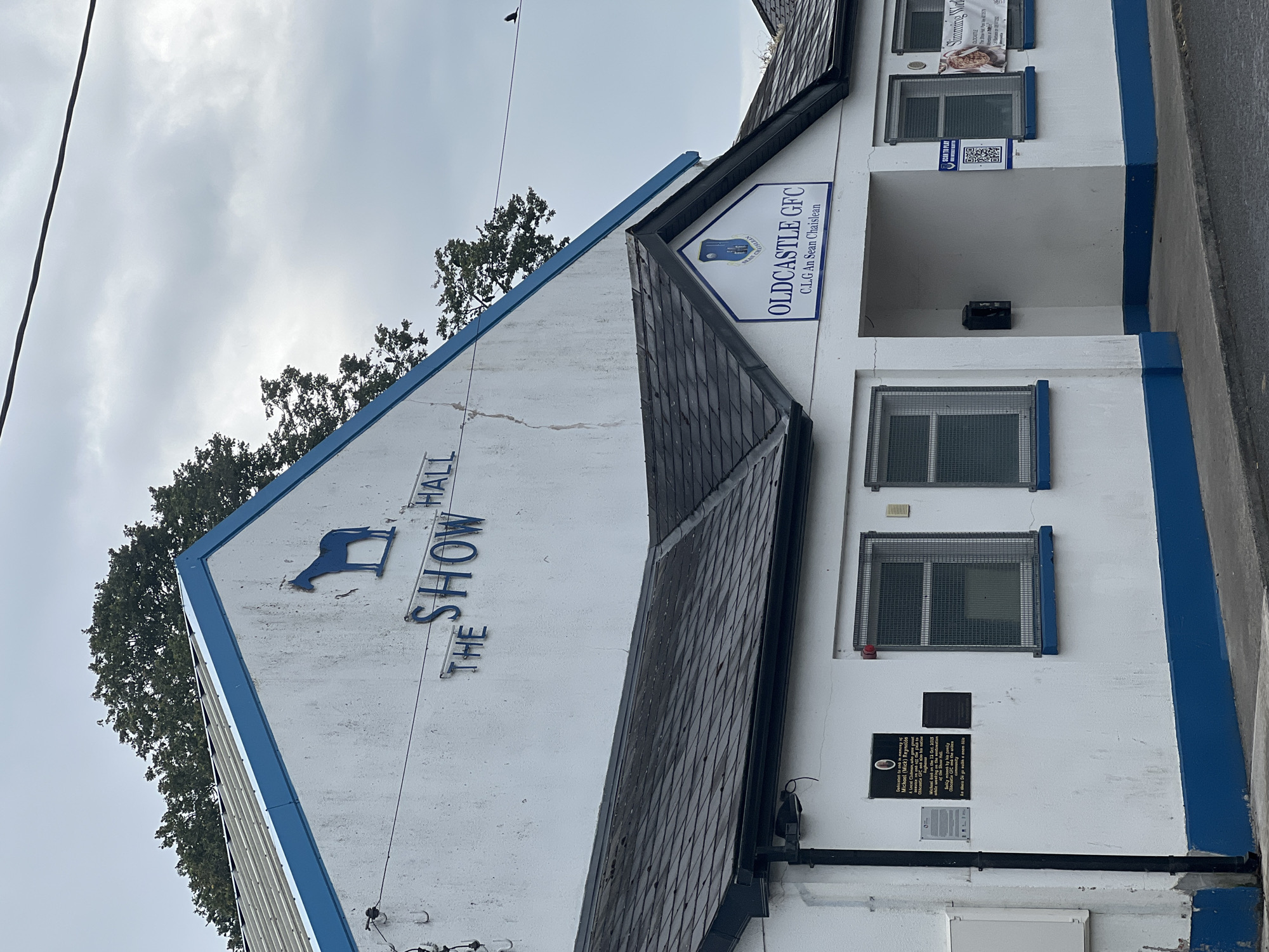 The image shows the exterior of a building with white walls and blue trim. The building has a pitched roof covered in dark grey shingles. On the upper portion of the building, a blue silhouette of a horse is mounted, and the words "THE SHOW HALL" are written in blue letters. Below this, a smaller section of the building has a sign with a shield logo and the text "OLDCASTLE GFC C.L.G An Sean Chaisleán". The building has several windows with metal grates. On the left side of the building, there are two plaques mounted on the wall. The environment appears to be overcast.