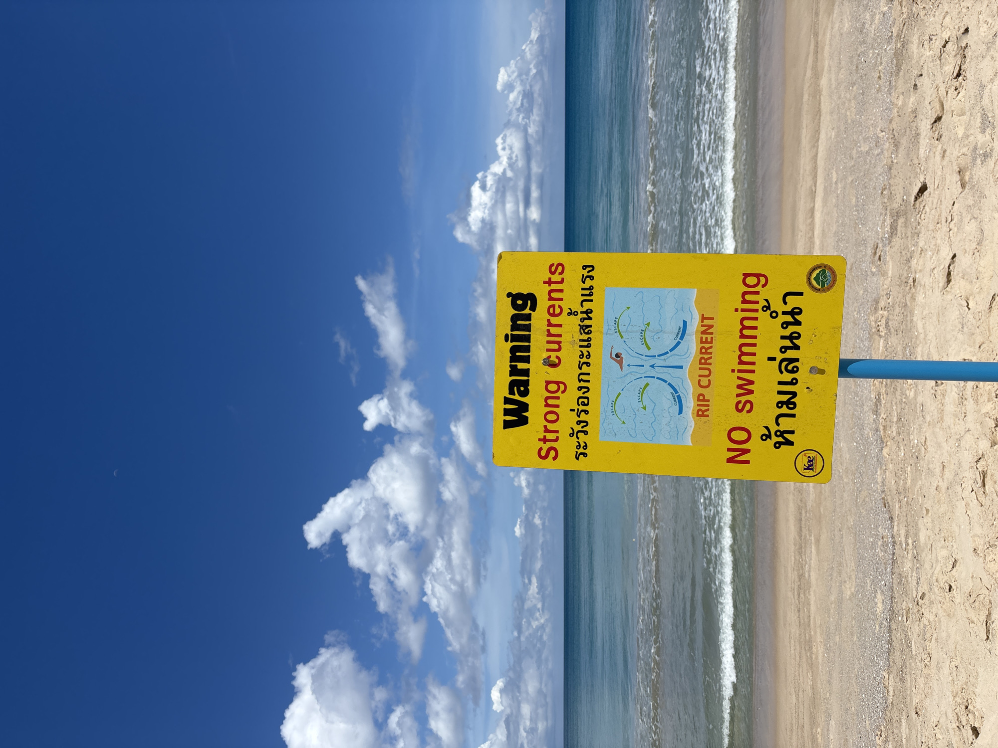 The image depicts a beach scene in Choeng Thale, Thailand. A yellow sign with English and Thai warnings about strong currents and the prohibition of swimming is prominently displayed. The sign features diagrams illustrating rip currents. The background shows the ocean with gentle waves meeting the sandy beach. Above, a bright blue sky is punctuated by fluffy white clouds.