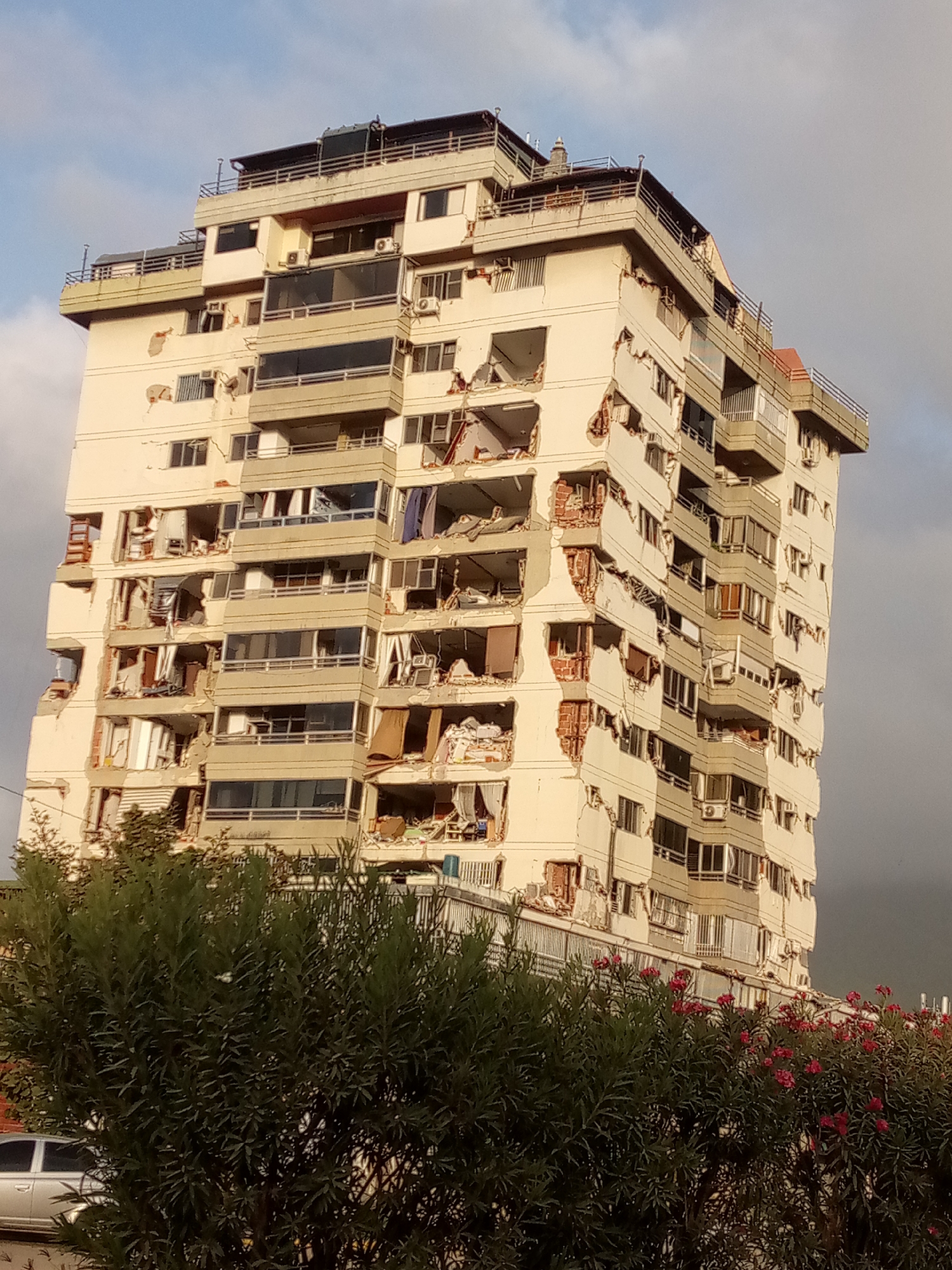The image shows a multi-story apartment building in Caraballeda, Venezuela, with significant structural damage. The building's exterior walls are cracked and have collapsed in multiple sections, revealing the interior of apartments, exposed brickwork, and debris. Many windows are broken, and curtains and other interior furnishings are visible. The building appears to have sustained damage from an earthquake or other catastrophic event. In the foreground, lush green foliage and pink flowers obscure the lower portion of the building and a silver car parked on the street. The sky above is a mix of blue with white clouds and darker, overcast areas. The overall scene depicts a residential area affected by a disaster.
