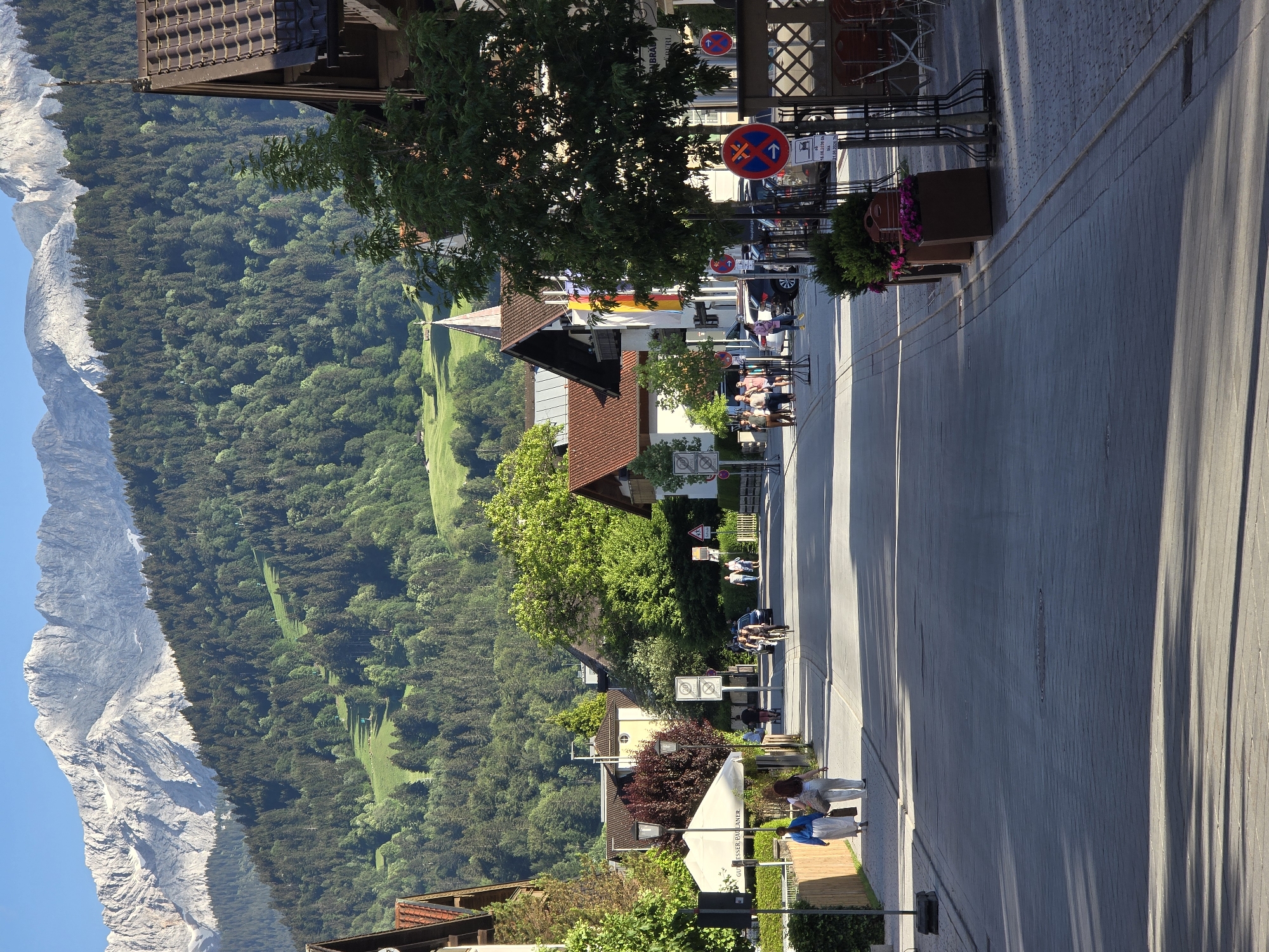 The image shows a street scene in Garmisch-Partenkirchen, Germany, with the Alps in the background. The street is paved with cobblestones and lined with buildings with traditional Bavarian architecture. There are several people walking on the sidewalk, and some parked cars on the street. In the foreground, there are some planters with flowers. The sun is shining, and the sky is clear. The mountains in the background are snow-capped, indicating it may be late spring or early autumn.