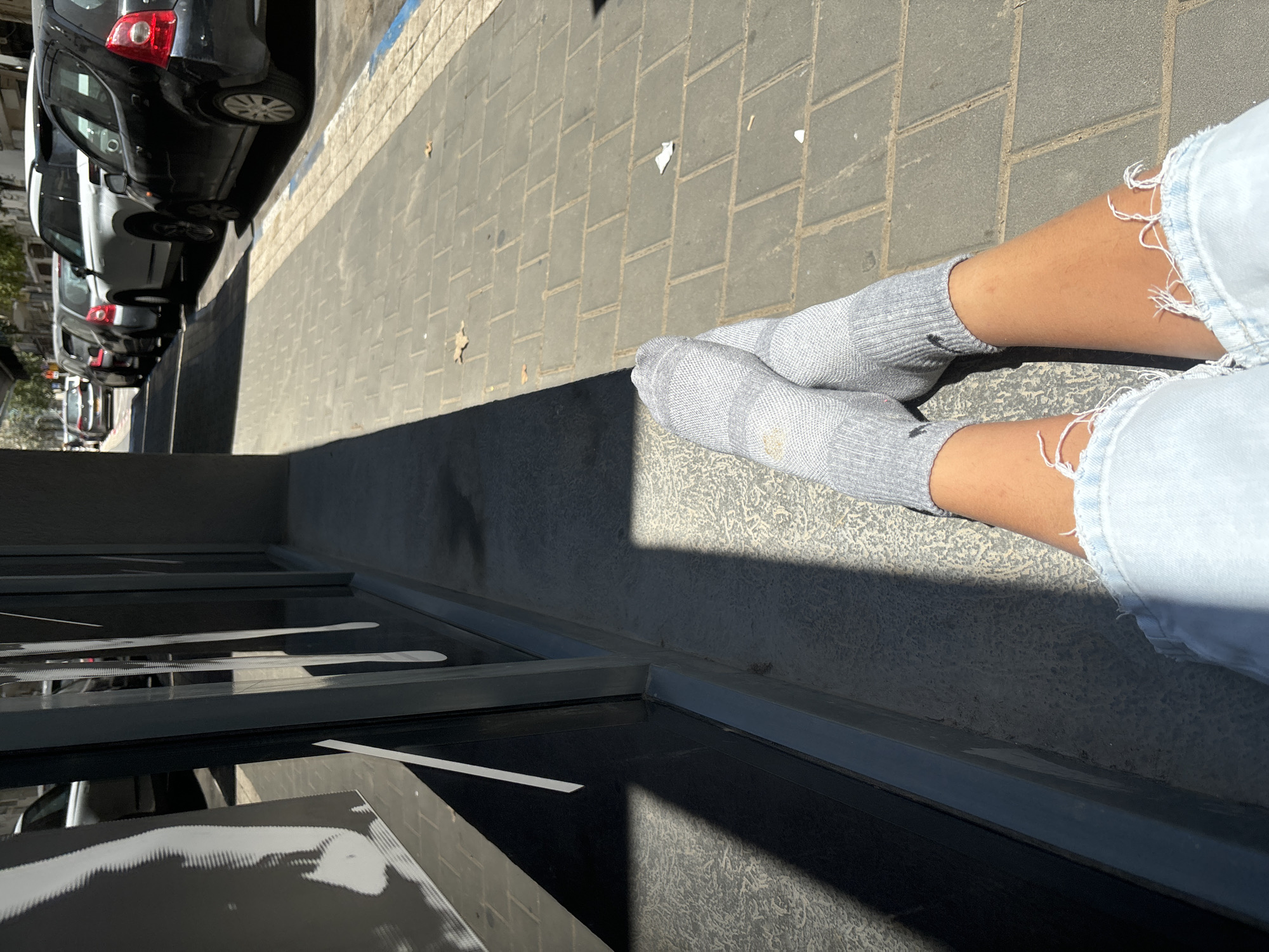 This image captures an outdoor, urban scene in Tel Aviv, Israel, from a low, slightly downward-angled perspective, seemingly from a seated position. In the immediate foreground, a dark, reflective surface, possibly a window or a building ledge, occupies the bottom and left side of the frame, showing blurred white reflections.
Dominating the right and center-right of the image are the lower legs of a person, extended onto a light gray paved sidewalk. The person is wearing light-wash ripped denim shorts or jeans, revealing tanned skin on their knees and lower thighs. Their feet are clad in light gray athletic socks, which bear a dark Nike swoosh logo on the outer side of each ankle. One sock shows a slight yellowish discoloration near the heel.
The sidewalk, made of large square paving stones, stretches across the mid-ground. Scattered small pieces of white paper and dried leaves are visible on its surface. Strong, bright sunlight casts sharp, dark shadows from the person's legs and other unseen elements onto the pavement and the reflective surface, indicating a clear, sunny day.
In the background, a row of parked cars, predominantly dark-colored compact vehicles, lines a street. A blue line painted on the curb suggests a designated parking area. The cars are parked parallel to the sidewalk, with some showing reflections on their bodies. The overall setting is characteristic of a bustling city street.