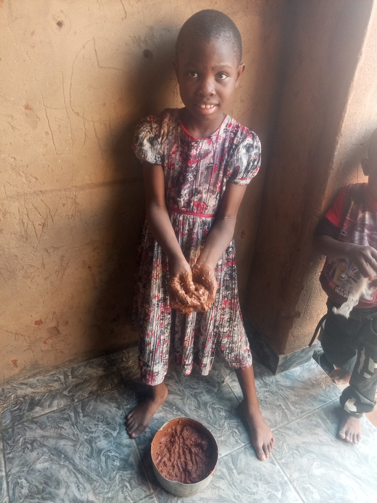 A young girl stands in a room. She is wearing a dress with short sleeves and a floral print. She holds a clump of brown matter in her hands. A metal bowl containing more of the same brown matter sits on the tiled floor near her feet. To her right, another child partially enters the frame. The child wears a red and black shirt and patterned pants. The walls of the room are roughly finished. The scene appears to be taking place in Tempure, Nigeria.