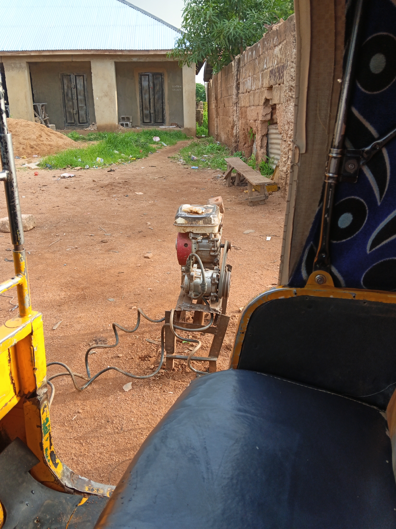 The image shows an outdoor scene in a dusty, rural environment. In the foreground, a portion of a yellow and blue vehicle, possibly a tuk-tuk or auto-rickshaw, is visible, with a dark blue upholstered seat. To the left, a yellow metal frame and part of a black floor mat are seen. In the center of the frame, a red and silver portable engine, likely a generator, is mounted on a metal stand. Black coiled hoses extend from the engine towards the vehicle.
The background features a light-colored building with a corrugated metal roof, two dark doors, and some greenery growing around it. A pile of sand is also present near the building. Further in the background, a weathered stone wall runs along the right side of the scene, with some vegetation growing through it. A simple wooden bench is placed against the wall. The ground is covered in reddish-brown dirt and scattered debris. The overall impression is of a utilitarian or working area, possibly in a developing region. There are no people visible in the image.