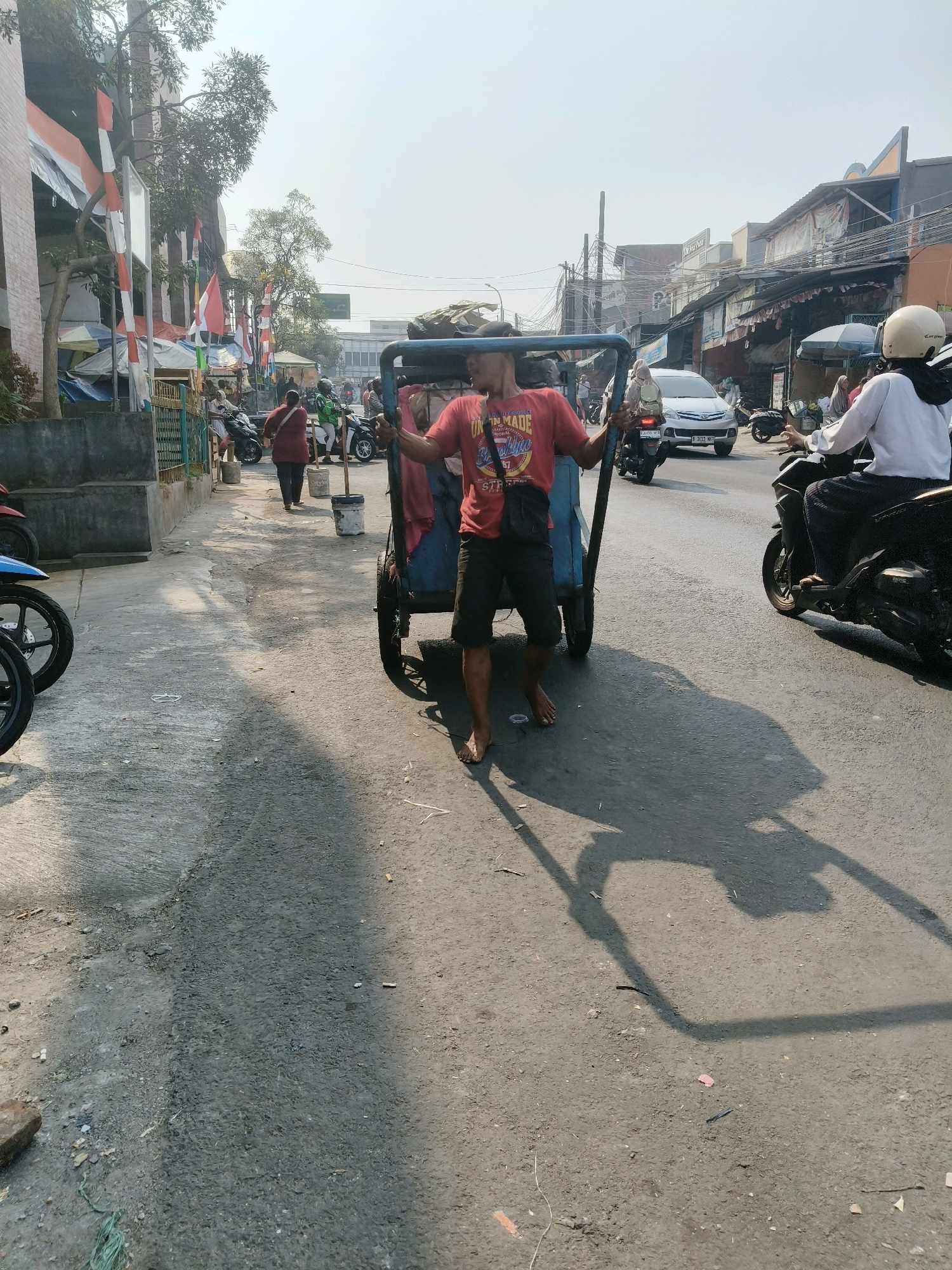 The image depicts a street scene in South Tangerang, Indonesia, on a sunny day with clear skies. A man in a red t-shirt and dark shorts is pushing a two-wheeled cart with a blue frame. The cart appears to be carrying some fabric or tarpaulin. The man is barefoot. To the right of the cart, a person wearing a white top and a white helmet is riding a motorcycle. In the background, there are various buildings, some with awnings and signs, suggesting a commercial area. Several other motorcycles are visible on the street, and some people are walking on the sidewalk. Indonesian flags are displayed on poles along the side of the road. The street surface is asphalt, with some debris scattered on it. The overall impression is of a bustling street during daylight hours.