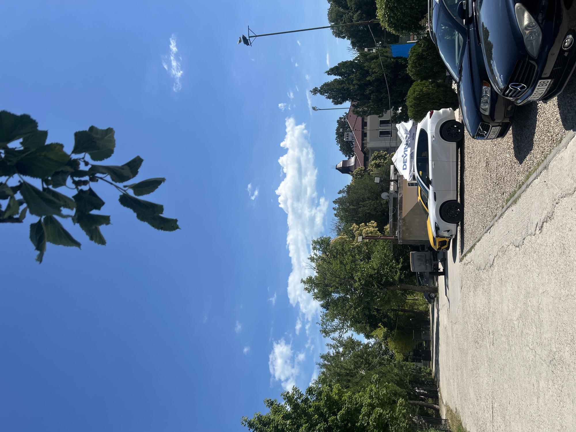 A sunny day is depicted in this outdoor shot, featuring a clear blue sky with scattered white clouds. The image is oriented vertically, with trees and buildings framing the lower half. In the foreground, a dark blue car is parked next to a white car with yellow accents. The white car appears to be a service vehicle, with the word "DEVIN" visible on its side, suggesting a bottled water company. A tent with the same branding is also present. The scene seems to be a parking lot or roadside area, with a paved surface and gravel. The lush green foliage of trees suggests a warm season. In the distance, residential buildings with tiled roofs and trees are visible. The time of day appears to be midday, given the bright sunlight and distinct shadows. No people are visible in the frame. The overall atmosphere is calm and serene.