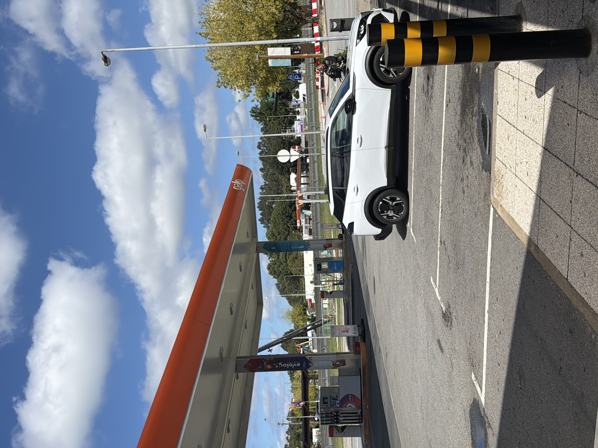 The image depicts a gas station in the city of Trofa, Portugal. A white SUV is parked at a pump under the orange awning of the gas station. The gas station has multiple pumps and signage, including a "Galp" logo. The ground is asphalt with white parking lines. Two black and yellow bollards are in the foreground. In the background, there are trees, streetlights, and other vehicles. The sky is blue with white clouds. A person is visible on a motorcycle to the right of the SUV.
