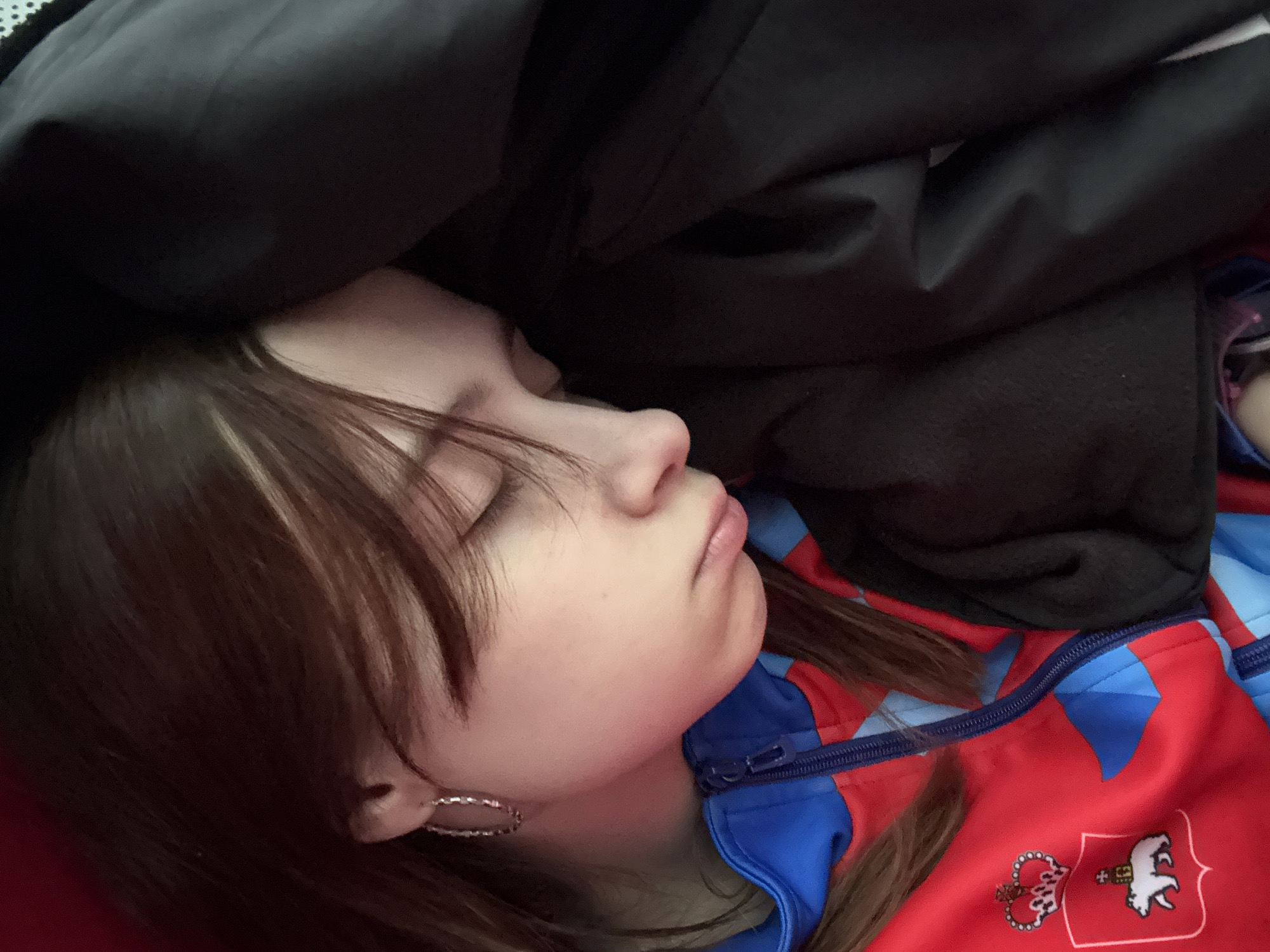 A close-up shot depicts a person sleeping. The person has fair skin, brown hair with bangs partially covering their face, and is wearing hoop earrings. They are dressed in a red and blue zippered jacket featuring a crest-like design with a white bear. A dark, seemingly heavy garment or blanket is draped over the person. The background is obscured, focusing attention on the sleeping individual. The location of Kukushtan, Russia, is not discernible from the visual information provided.