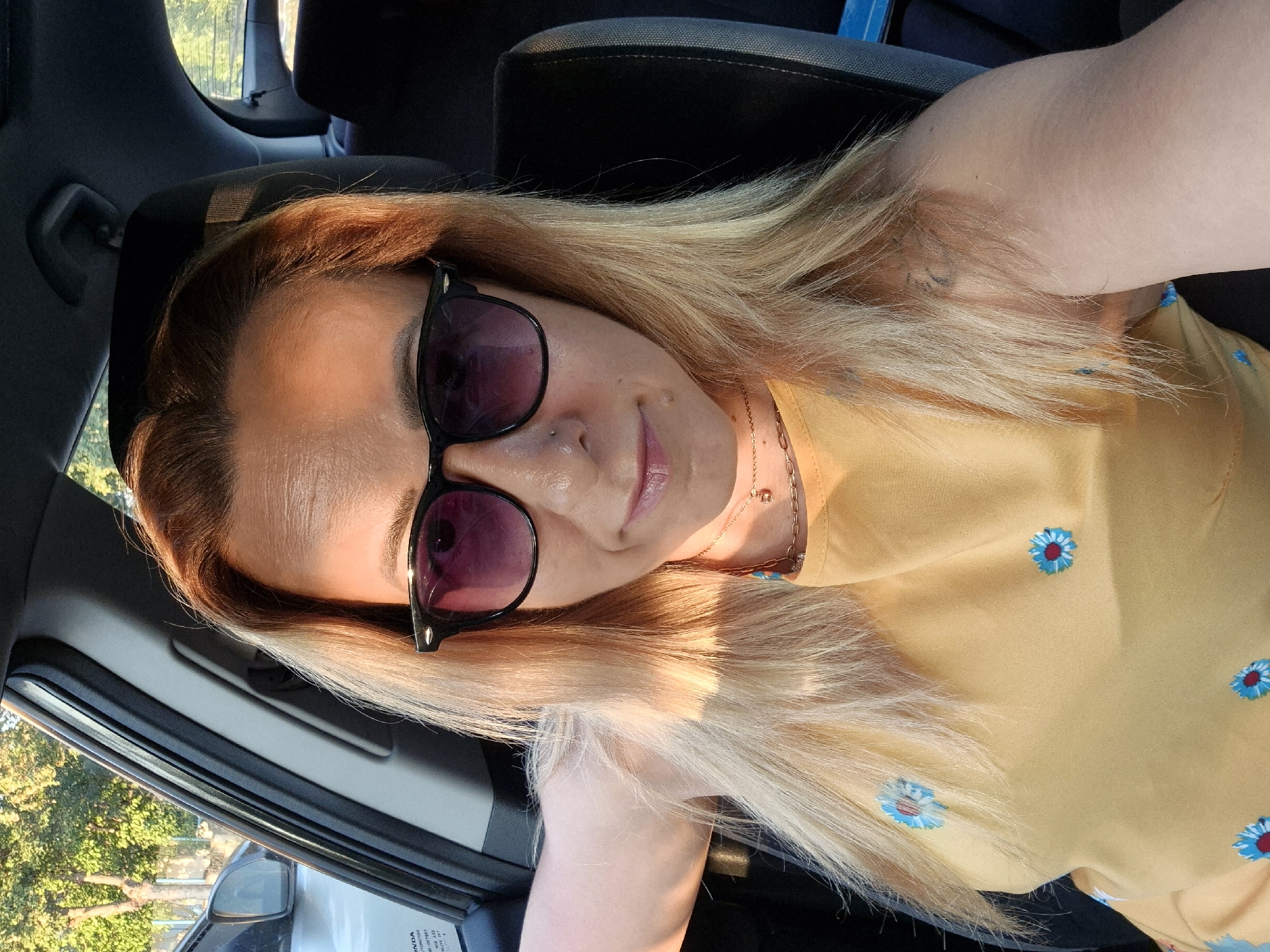 A blonde woman wearing sunglasses and a yellow top with blue flowers is sitting in the driver's seat of a car. She is looking at the camera and smiling slightly. The sun is shining on her face and hair. The background shows trees and a fence.