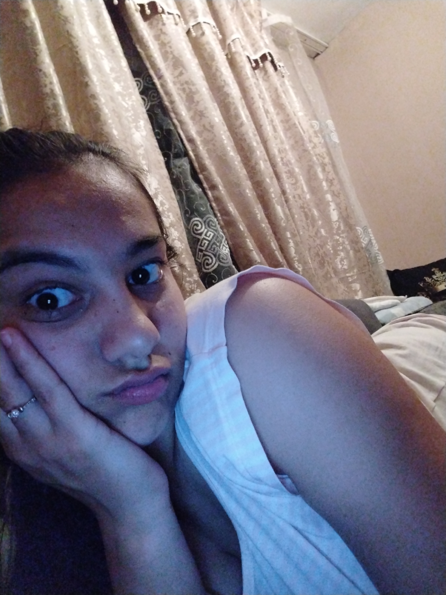 A close-up, slightly angled selfie shows a young woman with dark hair and eyes. She is lying down, resting her head on her left hand. A ring is visible on the ring finger of her left hand. She wears a light pink tank top. Behind her are draped curtains with a patterned design, and a portion of a wall. The specific location within the city of Unknown, Unknown is not discernible.