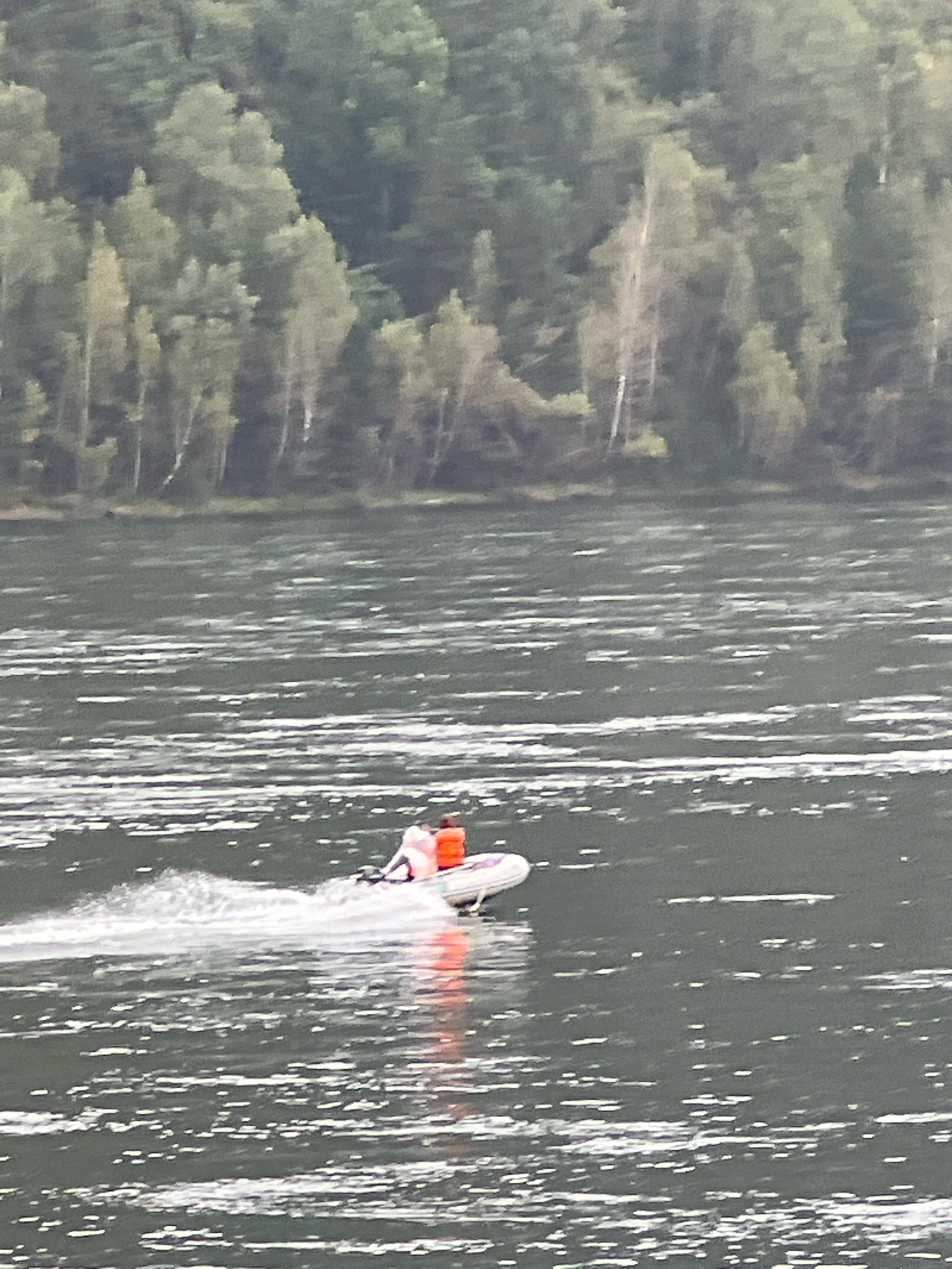 An inflatable boat with two occupants wearing orange life vests speeds across a body of water, leaving a white wake. The boat is moving from left to right. In the background, a densely wooded hillside lines the opposite shore of the water. The scene appears to be a natural landscape with no urban structures immediately visible, consistent with a location on the Krasnoyarsk Reservoir near Divnogorsk, Russia.