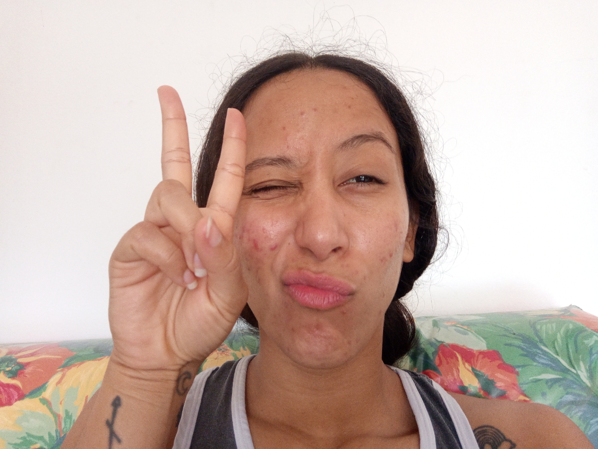 A medium shot captures a woman posing in front of a white wall with a floral-patterned surface behind her. She is fair-skinned and has dark hair pulled back from her face. Her skin shows some blemishes and marks. She is wearing a gray and white striped tank top.
The woman has her right eye closed in a wink, and her lips are puckered into a playful expression. Her right hand is raised, forming a peace sign with her index and middle fingers. A small tattoo is visible on her right wrist.
Another tattoo is visible on her left shoulder.
The setting might be indoors, given the plain white wall. The presence of the floral surface suggests a casual, possibly domestic environment.
This photograph could have been taken in Porlamar, Venezuela, or anywhere else, as the setting and subject don't provide definitive location clues.