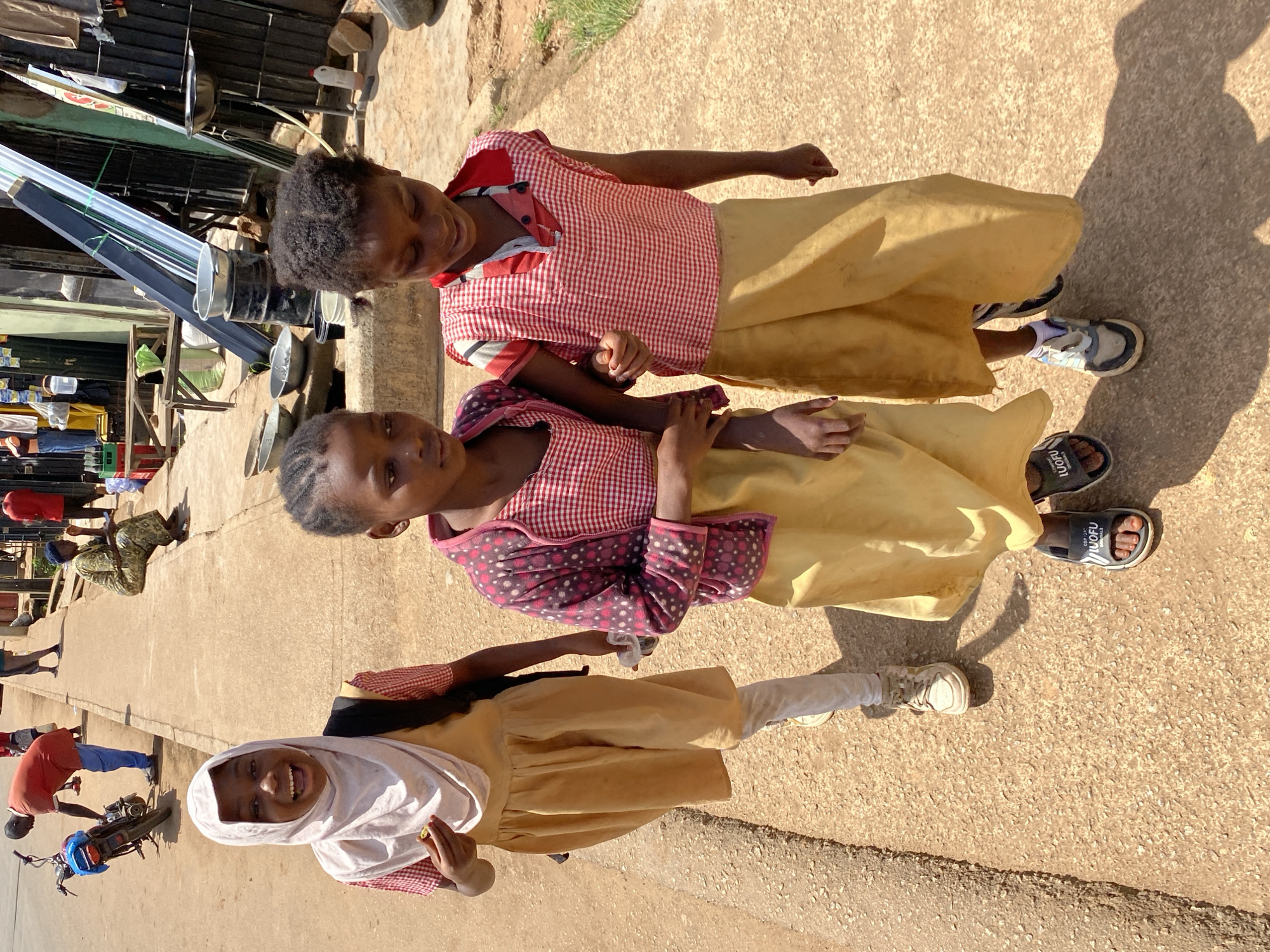 In Bukuru, Nigeria, on a sun-drenched, dusty street, three young girls walk together. The girl on the left, wearing a light pink hijab and a yellow dress with a red and white checkered top, beams a wide, joyous smile, holding a small yellow object in her hand. Next to her, the middle girl, also in a yellow dress and red and white checkered top, wears a patterned pink cardigan over it, her expression neutral as she walks. The third girl on the right, in a red and white checkered top and a yellow skirt, looks down, her arm linked with the middle girl's. Behind them, a busy street scene unfolds with a man in a red shirt and blue pants attending to a motorcycle, other people walking, and shops lining the street displaying various goods. The environment suggests a warm, dry climate with a ground surface of compacted earth and gravel.