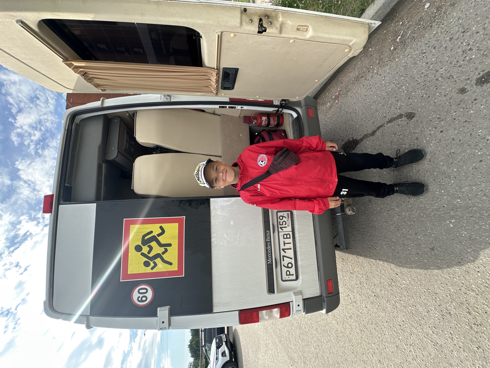 A young boy, wearing a red jacket, black pants, and a white baseball cap, stands in front of an open white Mercedes-Benz van. The van has a school bus sign with children running and a speed limit sign of 60 on its back. The license plate is visible and reads "P671TB 159 RUS." Inside the van, beige seats are visible, along with a red fire extinguisher and a red bag. The boy is smiling and looking at the camera. The scene appears to be on a paved road, possibly in a residential area, given the presence of trees and buildings in the background. The sky is cloudy. The van has "Mercedes-Benz" written on the back. The image is taken from a slightly low angle, looking up at the boy and the van.