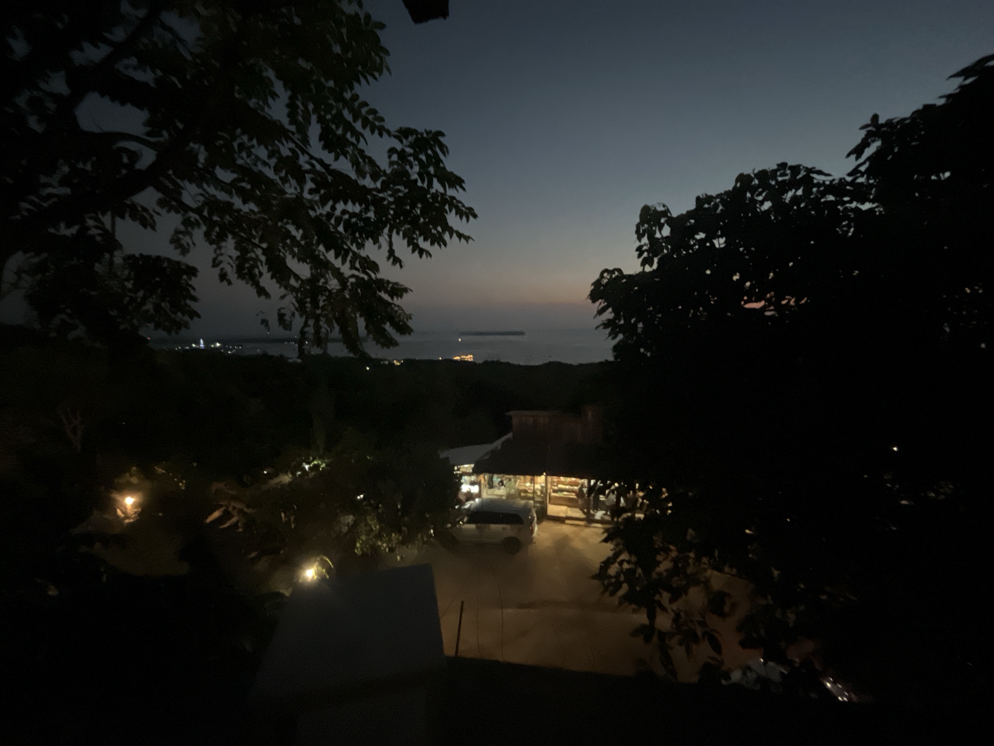 This image captures a twilight scene in Karimunjawa, Indonesia. The foreground is dominated by the dark silhouettes of trees and foliage, with a hint of a rooftop in the lower left corner. In the midground, a small building, possibly a shop or restaurant, is illuminated by warm lights, with a white van parked in front. Beyond this, the landscape stretches towards the sea, where a distant island or landmass is visible, dotted with lights. The sky transitions from a deep blue at the top to a lighter hue near the horizon, suggesting the sun has recently set or is about to rise. There are no discernible people in the image. The overall atmosphere is serene and quiet, characteristic of a peaceful evening in a coastal town.