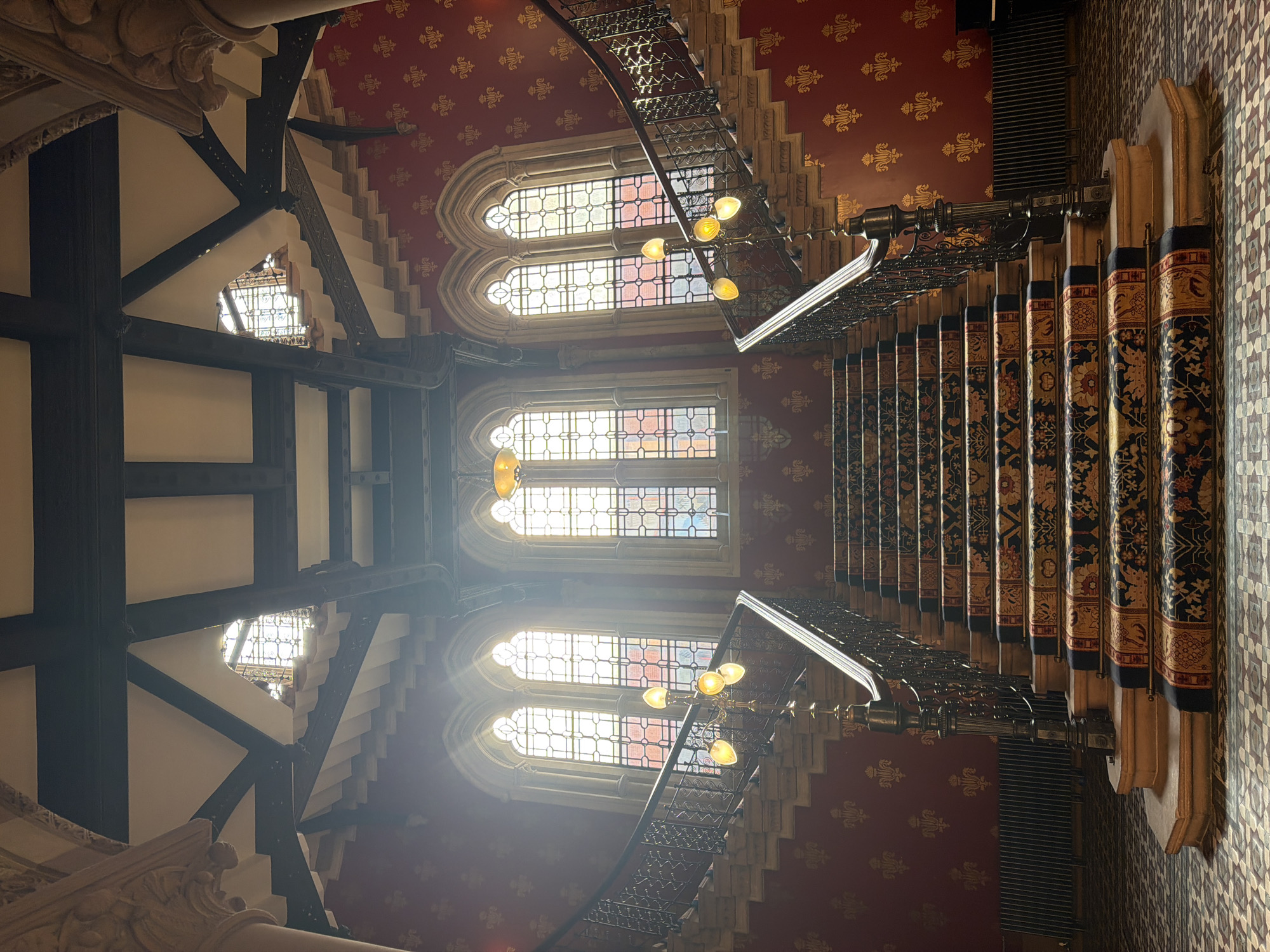 An opulent interior staircase is depicted, viewed from below and looking upwards through multiple levels of a grand, historic building, likely in London, United Kingdom. The image is oriented vertically, showing the height and intricate details of the architecture.
The prominent subject is a sweeping staircase with dark, ornate metal balustrades featuring detailed scrollwork. The steps are covered with rich, patterned carpet runners in dark blue or black, adorned with elaborate gold and red floral and geometric designs. The newel posts supporting the handrail are also intricately carved or cast.
The walls and high ceiling are covered in a striking deep red wallpaper featuring a repetitive pattern of gold heraldic motifs, possibly fleur-de-lis. Natural light streams in through several tall, arched windows with leaded or stained glass panes, some appearing to have muted colored sections, creating bright illumination and some lens flare. Ornate chandeliers with multiple bulbs hang from the ceiling, providing additional light.
Structural elements include dark, exposed wooden beams and decorative carved stonework or plaster detailing on columns and architectural supports, indicative of Gothic Revival or Victorian design. The scene is devoid of people. The bright light from the windows suggests it is daytime. No visible text is present in the image.