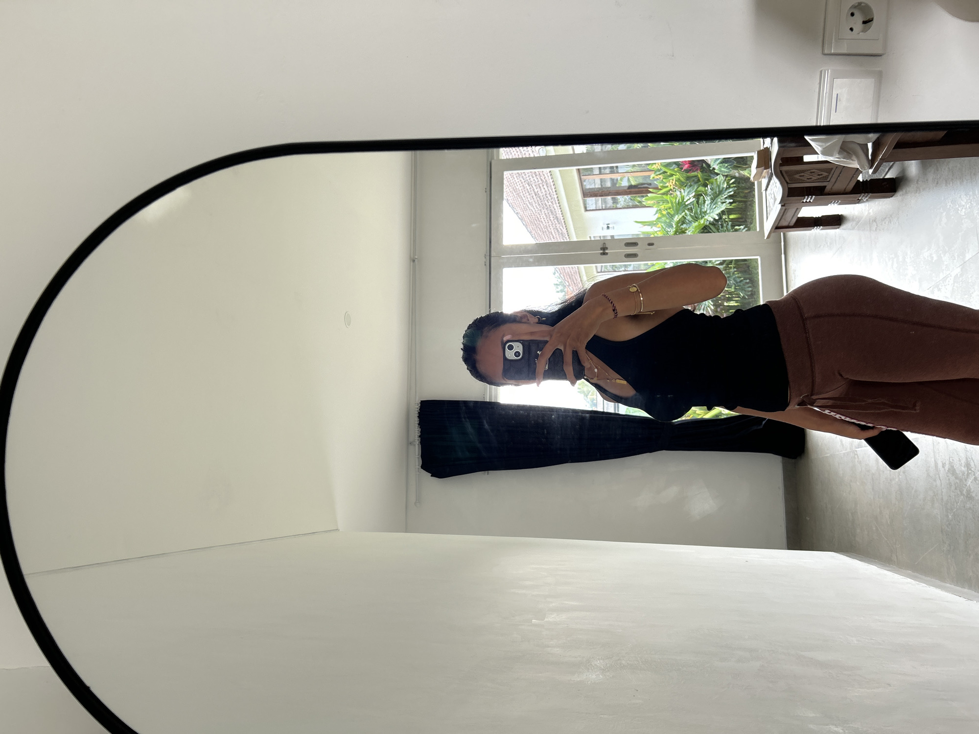 In Tembuku, Indonesia, a woman is taking a mirror selfie. She is wearing a black tank top and brown sweatpants. Her hair is dark and pulled back. She is holding a black smartphone in her right hand, which she is using to take the photo. The background shows a room with white walls and a large arched mirror. Through a set of glass doors, tropical foliage and a house can be seen. A wooden bench is placed in front of the doors. The floor is tiled.
