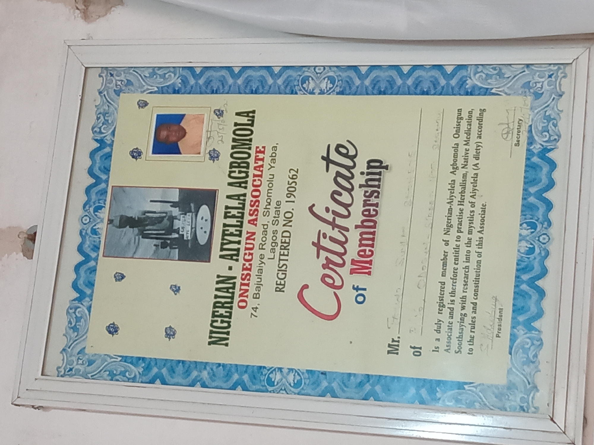 A framed "Certificate of Membership" is captured in a slightly tilted, close-up shot, displayed on a light-colored wall with some visible wear and tear. The certificate features a light yellow background with ornate blue borders.
The prominent text on the certificate identifies the issuing body as "NIGERIAN - AIYELELA AGBOMOLA ONISEGUN ASSOCIATE," located at "74, Bajulaiye Road, Shomolu Yaba, Lagos State," with "REGISTERED NO. 190562." The certificate attests that "Mr. Olabanji Sulaimon" is a registered member, entitled to practice "Herbalism, Native Medication, Soothsaying with research into the mystics of Aiyelela (A diety) according to the rules and constitution of this Associate."
Two small photographs are present on the certificate. The upper photograph is a color headshot of a dark-skinned man, likely Mr. Sulaimon, against a blue background, accompanied by a handwritten date "25/09/02." Below it, a black and white photograph depicts what appears to be a group of people or a scene, possibly related to the organization's practices or traditions.
The certificate bears signatures for the "President" (appearing as "Adegbang") and "Secretary." This document, signifying professional affiliation in traditional Nigerian practices, is displayed, indicating its importance to the individual, in Shagamu, Nigeria.