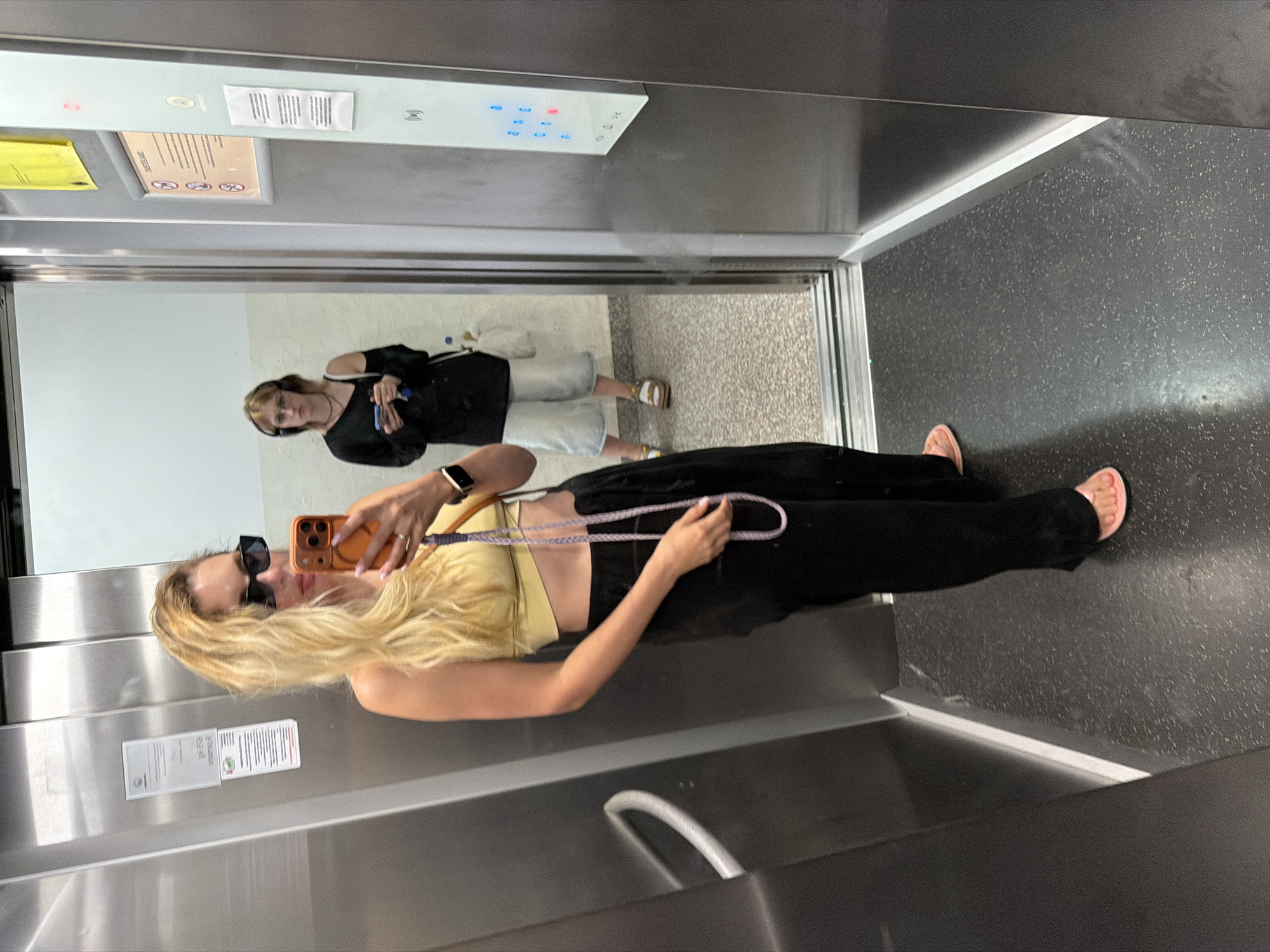 In an elevator, a young woman with blonde hair, wearing sunglasses, a yellow top, and black pants, takes a selfie. She holds an orange-cased phone in her right hand, and a braided strap hangs from her shoulder. In the mirror reflection, another woman with darker hair is visible, wearing a black top, denim shorts, and sandals, holding a blue object and a purse. The elevator appears modern, with stainless steel walls and a dark speckled floor. Above the reflection, elevator buttons are visible, including one illuminated in red. A sign with text and pictograms is affixed to the elevator wall. The scene suggests daytime, likely in a public building in Budva, Montenegro.