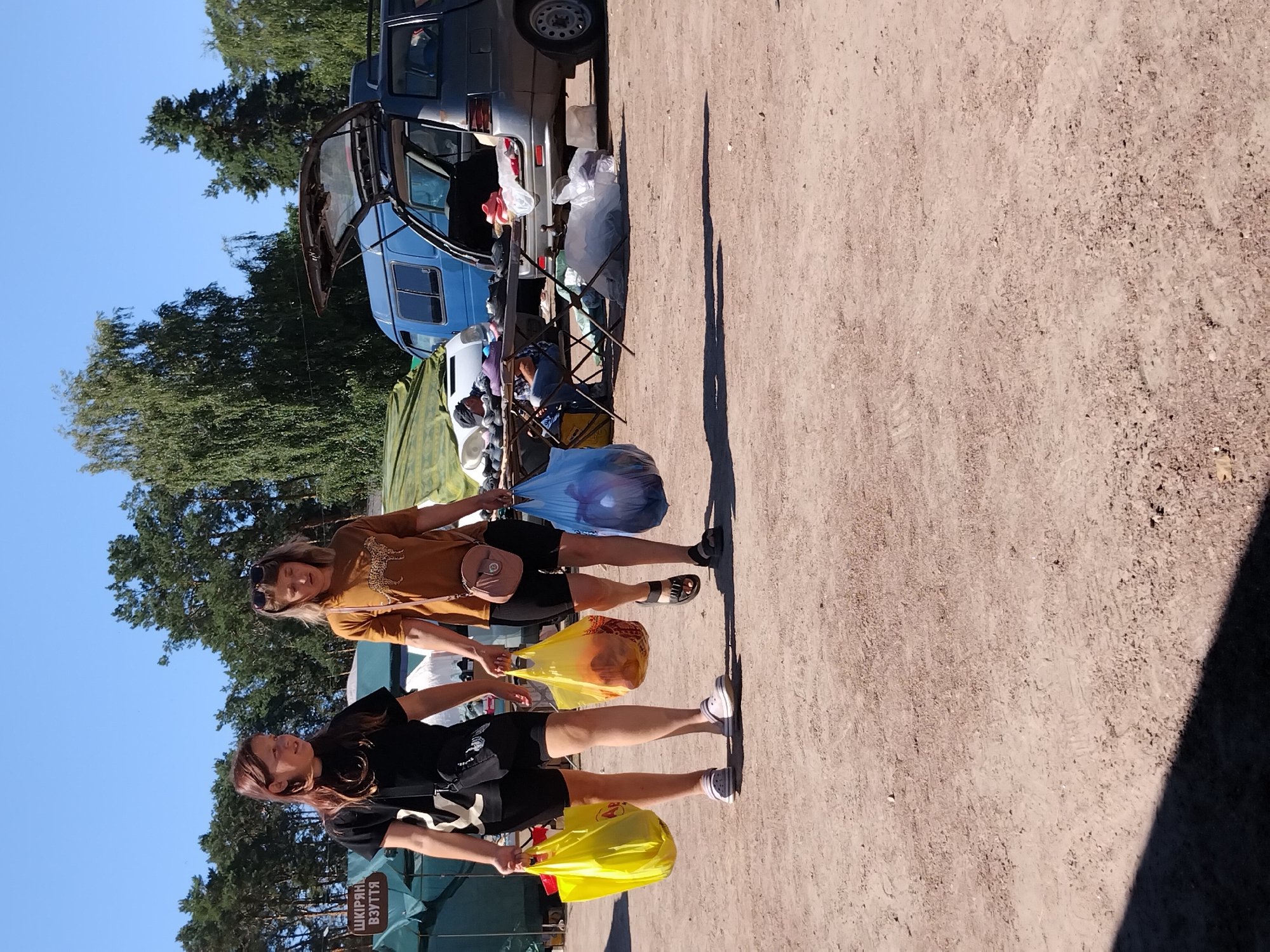 In Irshansk, Ukraine, two young women are walking outdoors, each carrying shopping bags. The woman on the left, with brown hair, wears a black t-shirt and black shorts, and carries a yellow shopping bag in her right hand. The woman on the right, with blonde hair, wears a tan t-shirt with a leopard print and black shorts, and carries a yellow shopping bag in her left hand and a blue shopping bag in her right. Both women appear to be in a parking lot or market area, with tents and vehicles in the background. The ground is dirt and gravel, and the sky is clear and blue.