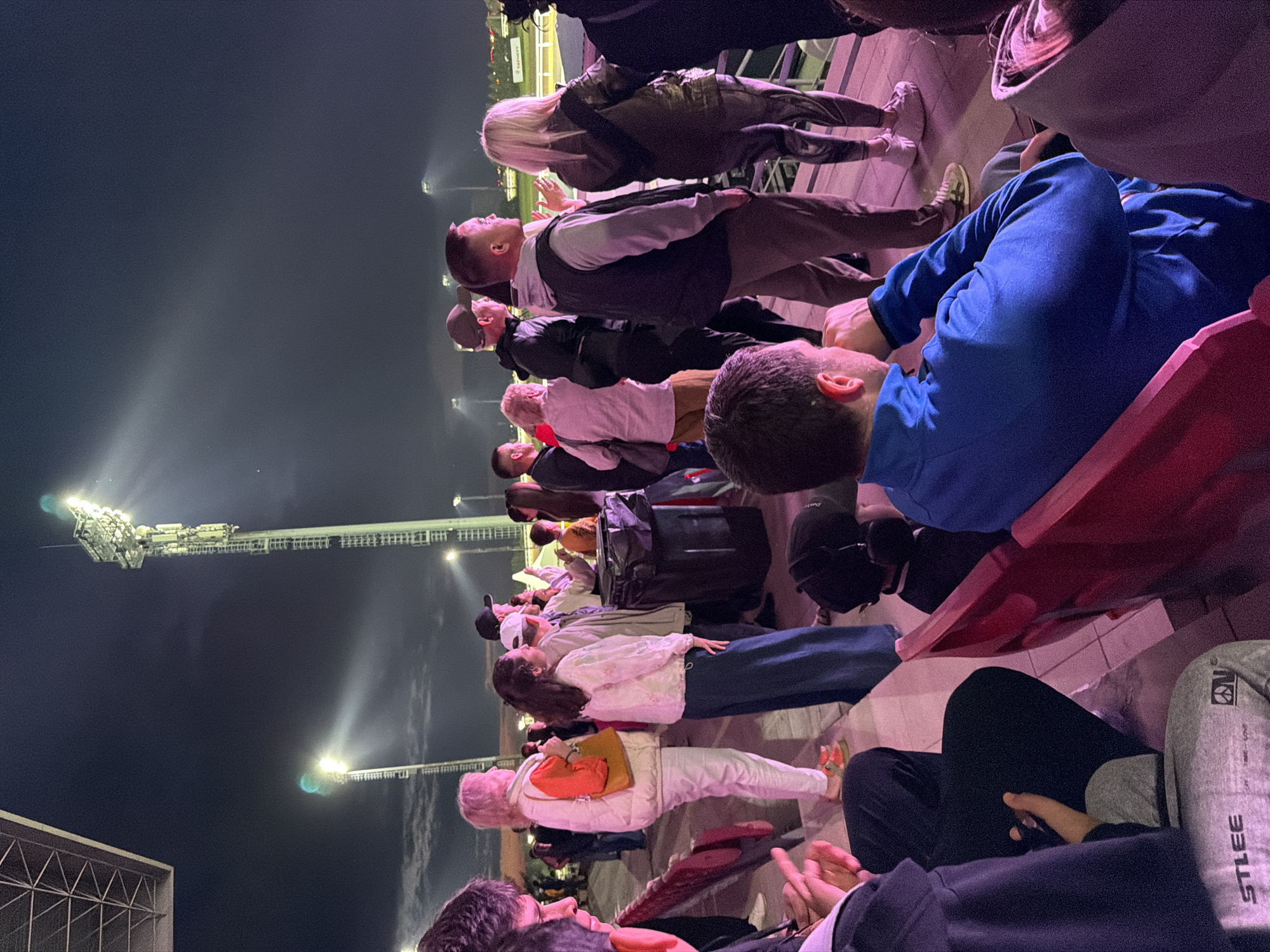 The image shows a crowd of people at an outdoor event at night. Several tall light towers illuminate the scene. In the foreground, people are seated and standing, some looking towards an unseen area. The people are dressed in casual clothing. In the middle ground, more people are visible, some in conversation. The background shows more light towers and a fence, suggesting a stadium or outdoor venue. The overall lighting is from artificial sources, creating a nighttime atmosphere. The location is described as Unknown, Unknown.