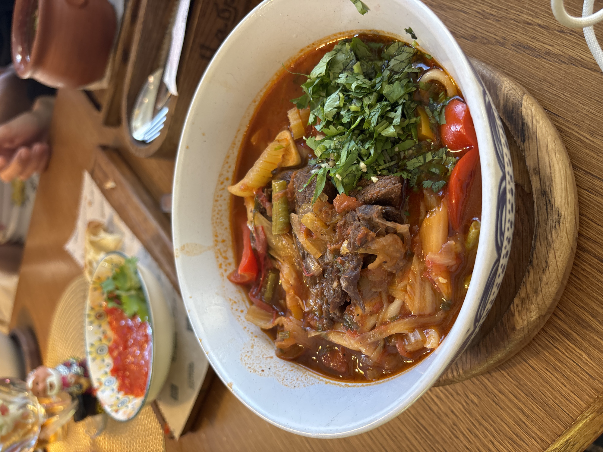 A top-down, slightly angled shot captures a bowl of hearty stew, likely a traditional dish from Solnechnoye, Russia, placed on a wooden table. The stew is rich with chunks of tender meat, visible green beans, and pieces of red bell pepper, all swimming in a savory, reddish-brown broth. The dish is generously garnished with finely chopped fresh green herbs, adding a vibrant contrast. The bowl itself is white with a decorative blue and white pattern along its rim, and it rests on a round, wooden serving board.
To the left of the main dish, a small, decorative ceramic bowl holds a bright red puree, possibly a sauce or relish, topped with a sprig of green herb. This smaller bowl also has a colorful, floral pattern. In the background, glimpses of other tableware and a hand reaching for something suggest a meal is in progress. The setting appears to be a restaurant or dining area, evidenced by the wooden table and cutlery holder in the upper left corner, which contains a spoon and fork. The lighting suggests it might be daytime. The overall impression is of a satisfying, home-style meal being enjoyed in a casual dining environment.