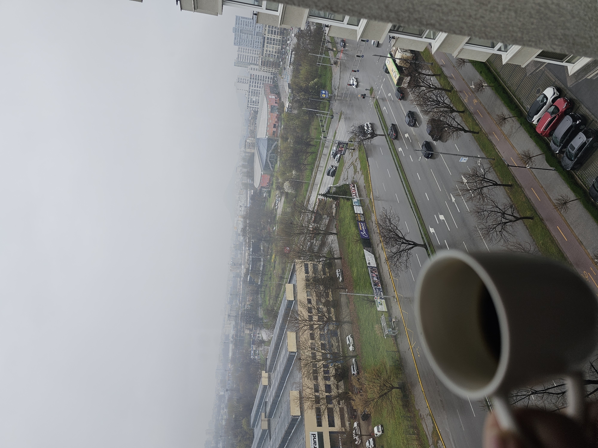 A high-angle, hazy daytime shot of a cityscape, viewed from a building. A cup of coffee is partially visible in the lower right corner. A multi-lane road bisects the scene, with cars moving in both directions. Parking on the building side of the road, where several vehicles are visible. Bare trees line the road. Buildings of various heights, including high-rise apartments and commercial structures, are located on either side of the road. A sports complex or arena can be seen in the distance. The weather conditions are overcast and foggy. This is in Plovdiv, Bulgaria.