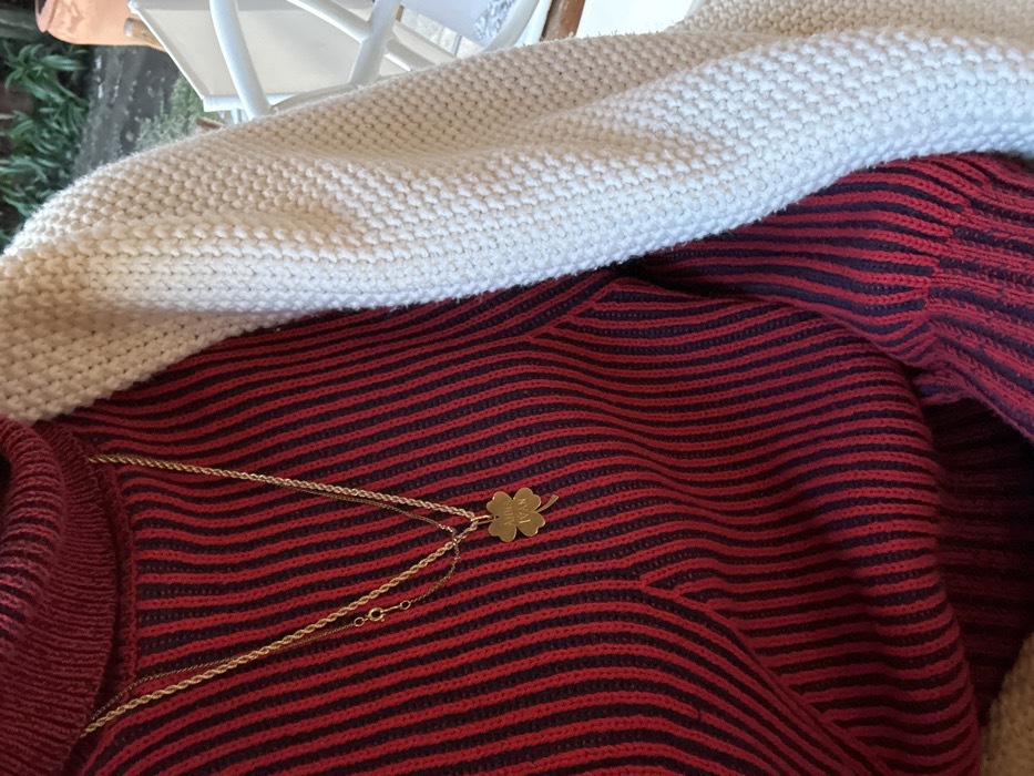 A close-up shot shows a gold necklace with a clover-shaped pendant resting on a red and black striped sweater. The clover charm appears to have writing or engravings on it. A thick, white knitted sweater is partially draped over the striped sweater, creating a layered effect. The garments are casually arranged, suggesting a relaxed or domestic setting.