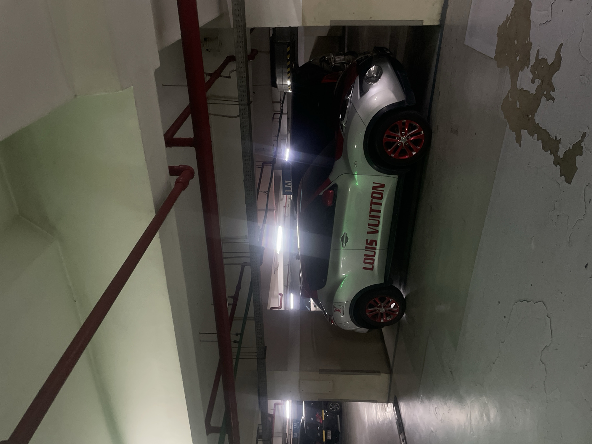 The image depicts a parked silver car with red accents and "LOUIS VUITTON" written in red across its side, situated in a dimly lit multi-story parking garage. The car is positioned in the foreground, partially obscuring a black SUV parked behind it. The garage environment is characterized by concrete pillars, exposed red pipes running along the ceiling, and overhead lighting fixtures that cast shadows. The floor of the garage appears to be concrete, with some water stains visible. In the background, other vehicles and structural elements of the parking garage are dimly illuminated. The scene is likely from a public parking area.