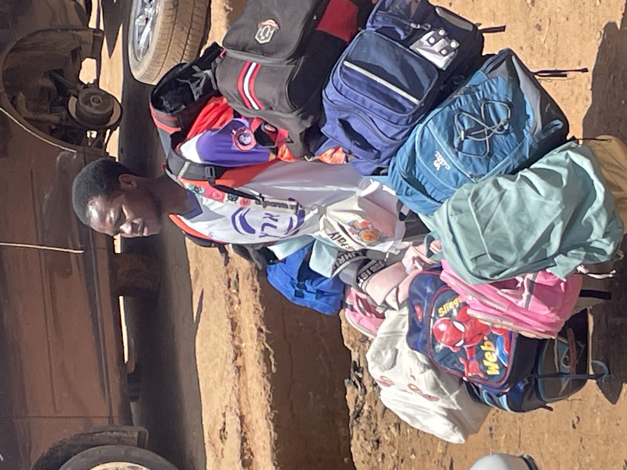 A young Black male stands outside, partially obscured by numerous backpacks. He is wearing a white t-shirt with purple and red accents, and a black strap is visible across his shoulders. The backpacks are piled high around him, in various colors including blue, black, pink, and white. One backpack features a Spider-Man graphic. The ground is a dusty, reddish-brown dirt. In the background, a dark-colored car with a missing front wheel and a detached tire resting nearby is parked on the uneven terrain. The overall scene suggests a street or roadside environment in Jos, Nigeria, possibly a transportation hub or market area given the loaded bags.