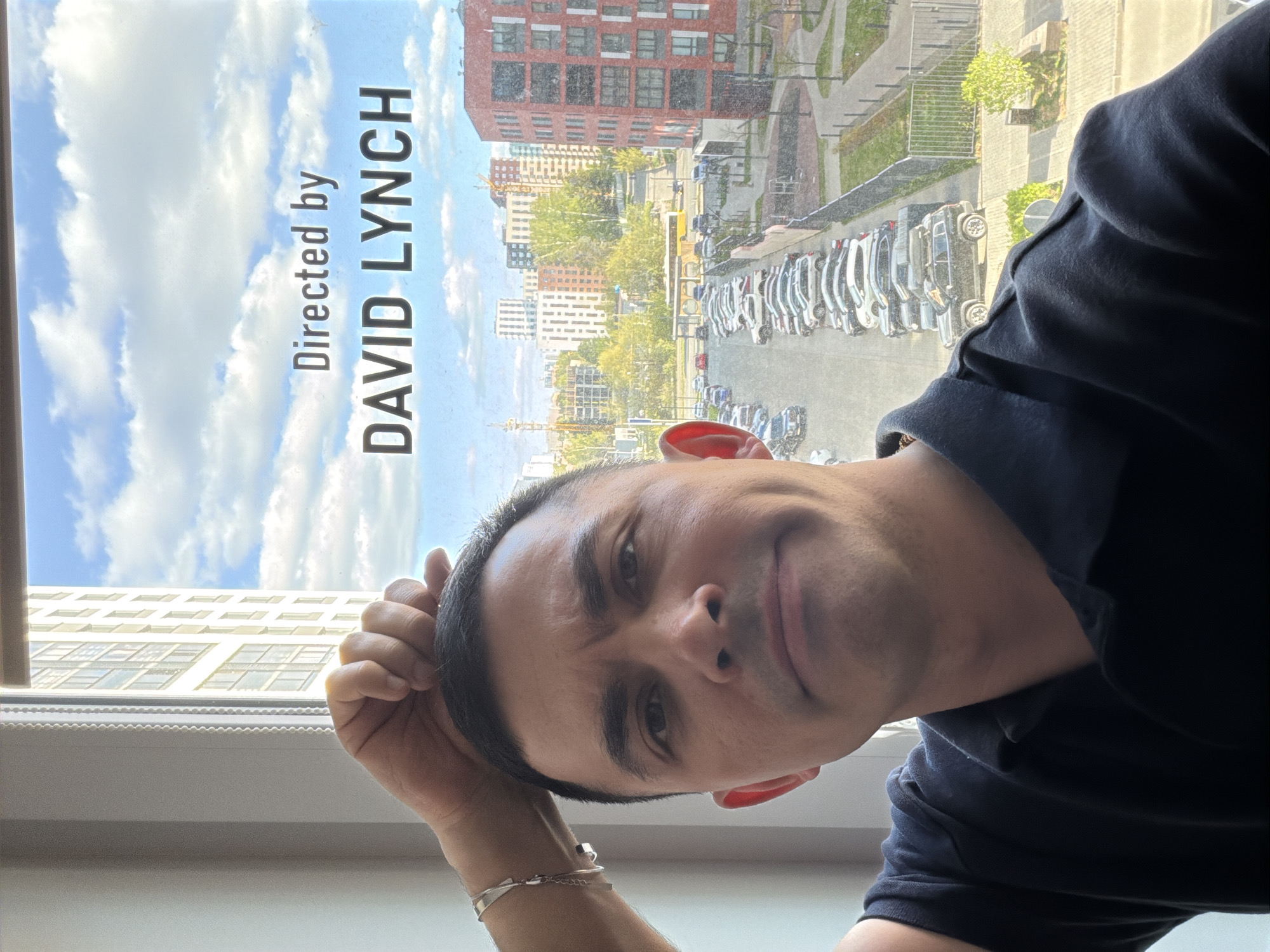 A man poses for a selfie indoors. The background shows a view through a window, featuring a city scene. A poster or picture in the window displays the words "Directed by DAVID LYNCH." Outside the window, buildings of various heights and colors are visible. A parking lot filled with cars is also present.
Based on the architectural style and visible surroundings, it is highly probable the image was taken in Izhevsk, Russia. The building architecture and parking lot are consistent with urban areas in Izhevsk.