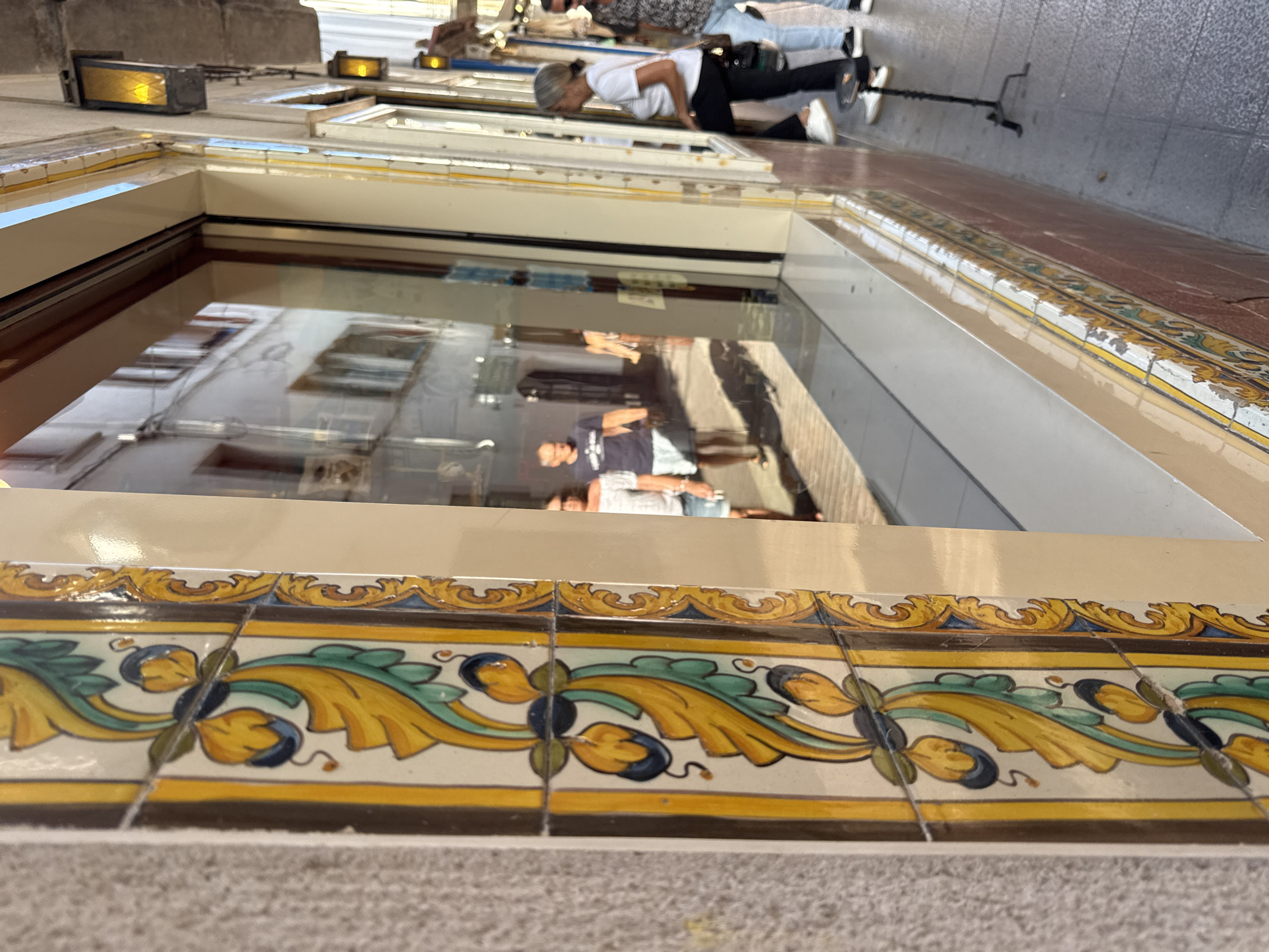 The image shows a reflection in a large window, revealing people and buildings. The reflection is distorted and upside down, suggesting the photo was taken from a low angle looking upwards. In the foreground, decorative tiles are visible, featuring a yellow, green, and blue floral pattern. These tiles appear to be on the exterior of a building.
The reflection shows several individuals. One person, in the reflected image, is wearing a white t-shirt and black pants, and is in a bent-over position, possibly looking into something. Another person in the reflection is wearing a dark t-shirt and shorts. Beyond them, more people and the facades of buildings are visible, suggesting an urban or town setting. The lighting in the reflection suggests it is daytime. The presence of decorative tiles and the architecture hint at a location in Spain, consistent with the provided context of Tossa De Mar. The overall scene appears to be a street view captured through a reflective surface, with the focus on the details of the building's exterior and the inverted reflections of the activity happening in front of it.