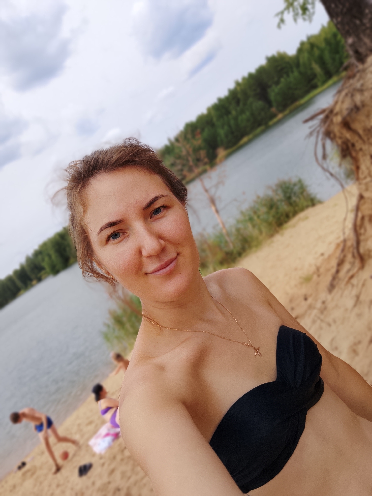 The image shows a woman taking a selfie on a sandy beach next to a lake. She is wearing a black bikini top and a gold necklace. Her hair is light brown and pulled back. She has blue eyes and is smiling.
In the background, the lake is calm and reflects the cloudy sky. Trees line the opposite shore, creating a natural backdrop. There are a few other people on the beach, including a child playing with a ball and a woman in a purple bikini sitting on a towel. The ground is covered in sand, and tree roots are visible on the right side of the image, suggesting the beach is near a wooded area.
The weather appears to be overcast, as indicated by the clouds in the sky. The overall mood of the image is relaxed and casual, suggesting a summer day spent outdoors. There is no visible text in the image. The setting is likely a natural recreational area in Noviy, Russia, given the presence of a lake and forest.