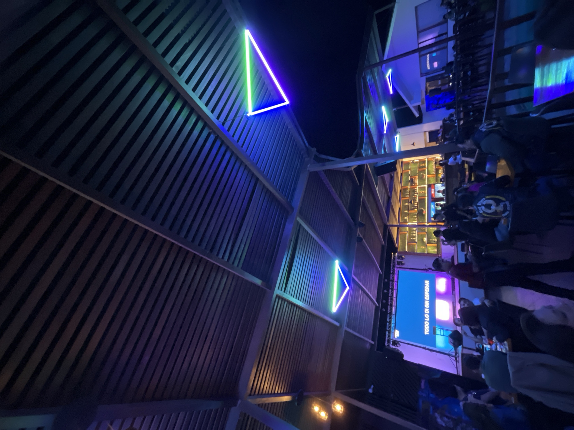 The image depicts an indoor event or gathering in a bar or restaurant, potentially in Arica, Chile. The scene features a crowd of people, possibly patrons, and staff members inside a multi-level establishment. A large screen displays the words "TODO LO DI SIN ESPERAR", suggesting an event of some kind.
The architecture features angled wooden slats on the ceiling, adorned with neon triangle lights, primarily in blue and green. The bar area is visibly stocked, and a possible upper level with a railing and more people can be seen.
Details about people are obscured, but various individuals are seen standing, interacting, and engaging in the event. The event appears to be a social gathering taking place at night.