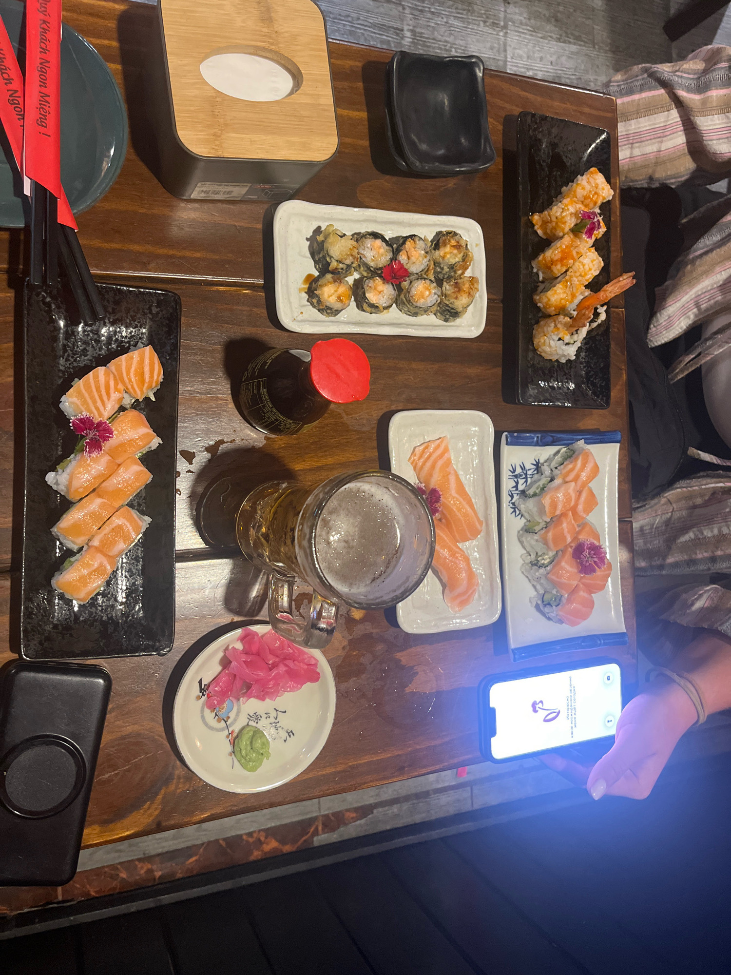 A top-down, eye-level shot shows a wooden table laden with various plates of sushi and a glass of beer. The scene is indoors, with a subtle brick-patterned wall visible in the background. Chopsticks rest in a black holder to the left, next to a dark green plate and red paper chopsticks sleeves with Vietnamese text that translates to "Wish you a delicious meal!".
To the right of the chopsticks, a light-colored rectangular plate holds several pieces of deep-fried sushi, garnished with red edible flowers. A small bottle of soy sauce with a red cap sits nearby. A large beer mug, half-full with frothy beer, is positioned below the soy sauce bottle. Next to the beer is a white oval dish containing bright pink pickled ginger and a small dollop of green wasabi.
Further to the right, a long, dark rectangular plate displays six pieces of sushi topped with salmon and a small purple flower. Adjacent to this, a white rectangular plate with blue trim holds a serving of sushi. Above these plates, a small black dish is empty. In the lower right corner, a hand is holding a smartphone, its screen illuminated with a flower graphic and Vietnamese text. A striped blanket or fabric is draped over a person to the right, partially obscuring them. The setting appears to be a restaurant in Nha Trang, Vietnam, given the Vietnamese text on the chopsticks.