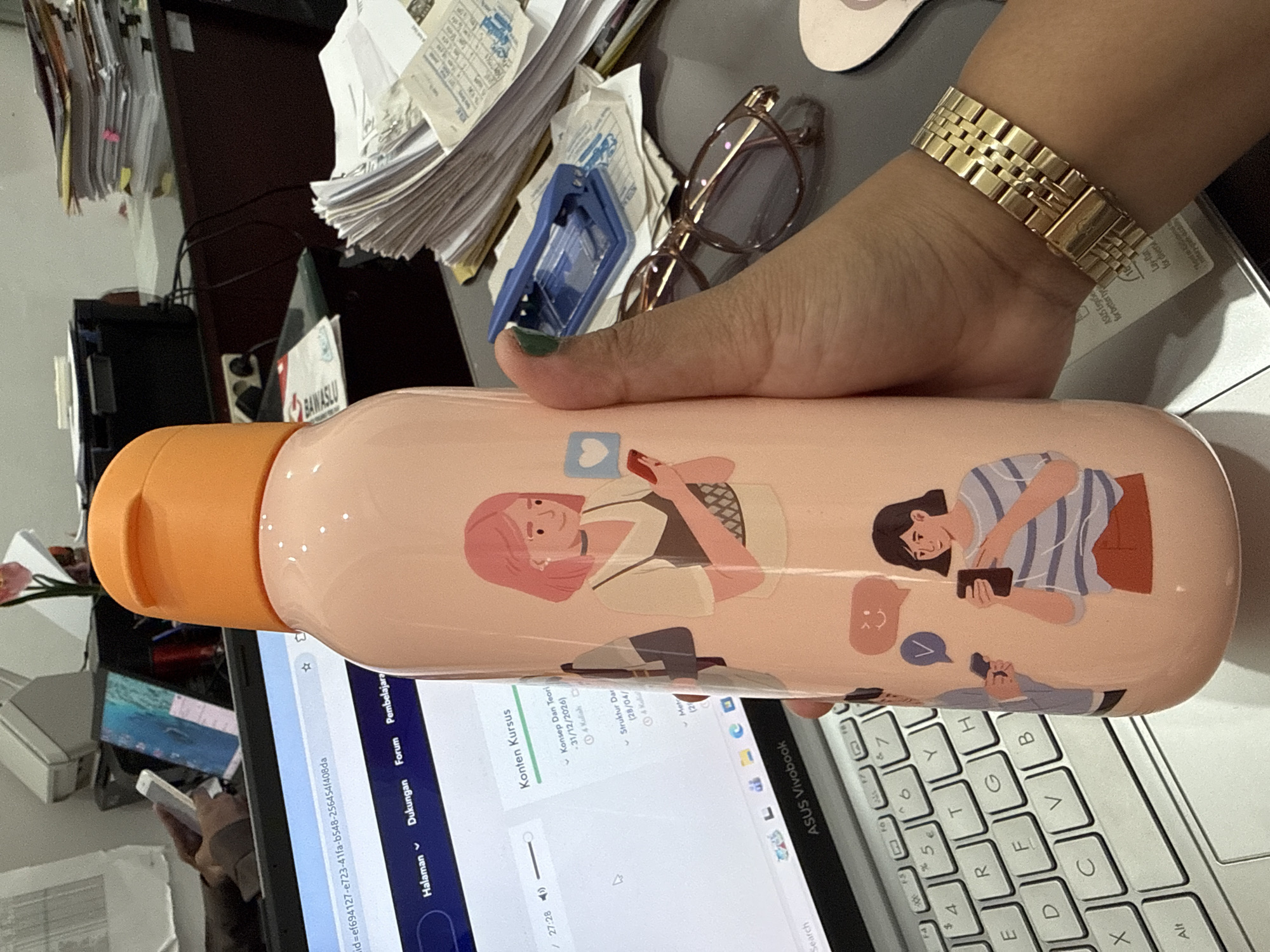 The image shows a person holding a pink water bottle with illustrations of people using smartphones. The bottle is placed on a laptop keyboard, specifically an ASUS Vivobook. The laptop screen displays a website or application with Indonesian text, indicating that the content includes "Konten Kursus" (Course Content), "Halaman" (Page), "Dukungan" (Support), "Forum," and "Pembelajara" (Learning). The website also shows dates "31/12/2026" and "28/04."
The person holding the bottle is wearing a gold watch. Behind the laptop, there's a messy desk with piles of papers, a pair of glasses, and other office supplies. The background is cluttered, suggesting a busy workspace.
The OCR text on the paper is likely the Indonesian Election Supervisory Body, "BAWASLU."
Given the language on the laptop screen and the likely presence of Indonesian documents in the background, the scene is probably located in Indonesia. However, there is no indication of it specifically being in Lhokseumawe.