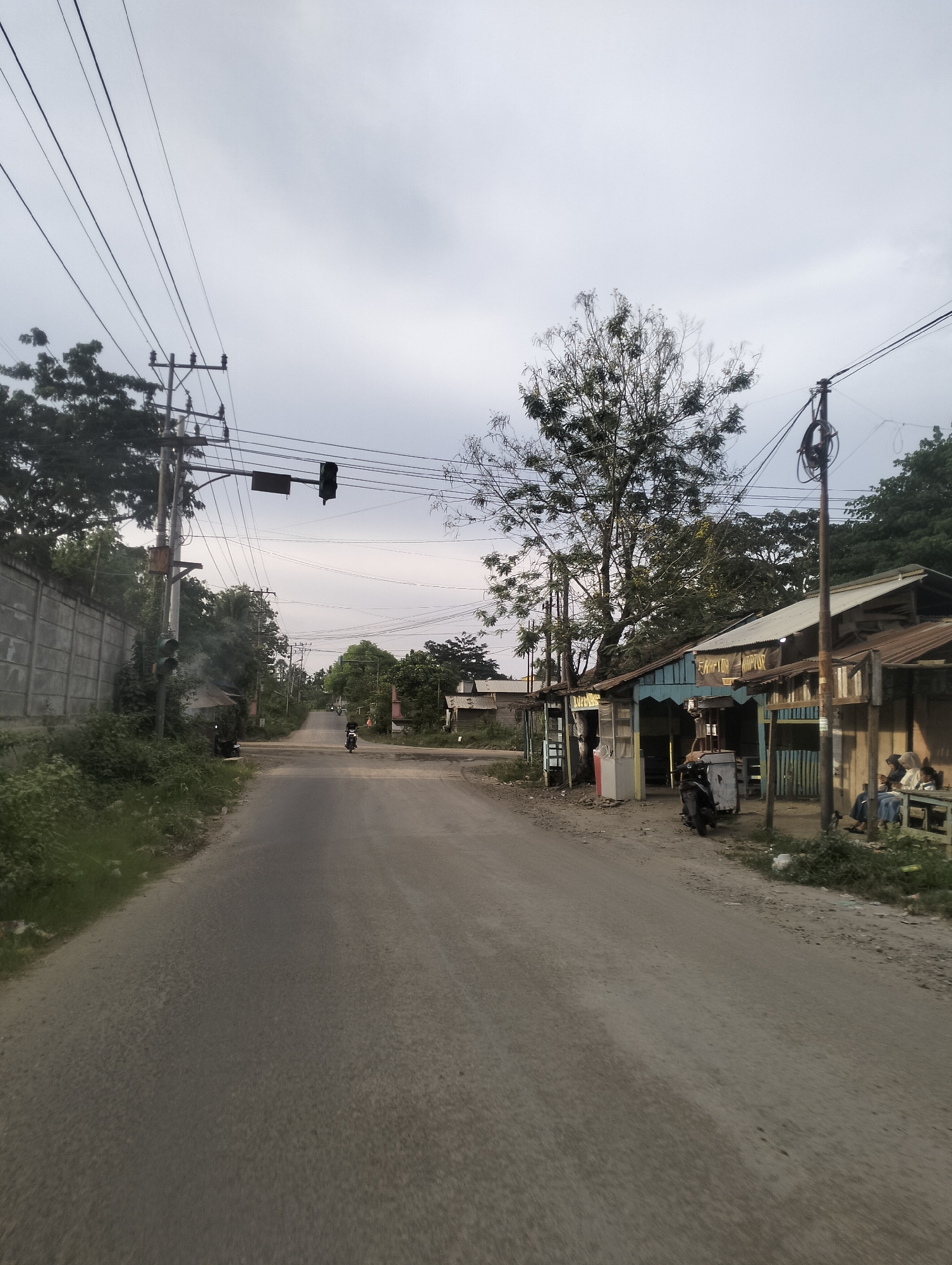 The image depicts a street scene in Lhokseumawe, Indonesia, likely during the late afternoon or early evening, given the overcast sky and diffused lighting. The setting is a rural or semi-urban road, characterized by a paved asphalt surface. On the left side of the road, a concrete wall separates the street from a lush, green area with dense vegetation. Electrical poles with multiple wires stretch across the sky, indicating a well-connected area despite the rustic appearance of the buildings.
Dominating the scene on the right are several modest structures that appear to be small shops or food stalls. These buildings have corrugated metal roofs and open fronts, with some displaying signage. One sign, partially visible, reads "KAPIE." Another sign above a stall reads "FAHRIY KOPTOR." These suggest commercial activity. Parked near these stalls is a motorcycle, and further down the road, another motorcycle is being ridden by a single person. A few individuals are seated on a bench outside one of the shops, seemingly socializing or observing the street.
The overall atmosphere is calm and ordinary, with no specific event prominently unfolding. The sky is uniformly gray, suggesting cloudy weather, but there's no indication of rain. The presence of traffic lights at an intersection further up the road, though partially obscured, hints at a functional roadway. The scene conveys a sense of everyday life in a community, with small businesses operating along the roadside.