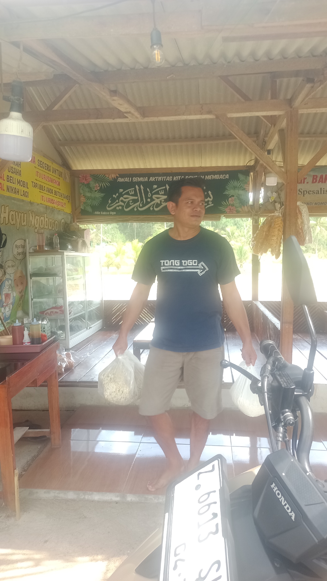 A man stands in what appears to be an outdoor food stall or small shop in Cibeber, Indonesia. He is wearing a dark blue t-shirt with the text "TONG DGC" and a white arrow pointing to the right, and tan shorts. He is holding a clear plastic bag filled with a light-colored, granular substance in his left hand and another similar bag in his right hand. The background shows a wooden structure with a corrugated metal roof and some plants visible through an opening. There are banners with text in Indonesian and Arabic script. To the left of the man is a wooden table with condiments and a glass display case filled with items. To the right, a portion of a motorcycle is visible, including its handlebars and front fender, with the "HONDA" logo clearly displayed. The floor is tiled with brown and beige tiles, and there is some exposed concrete. The overall lighting suggests it is daytime.