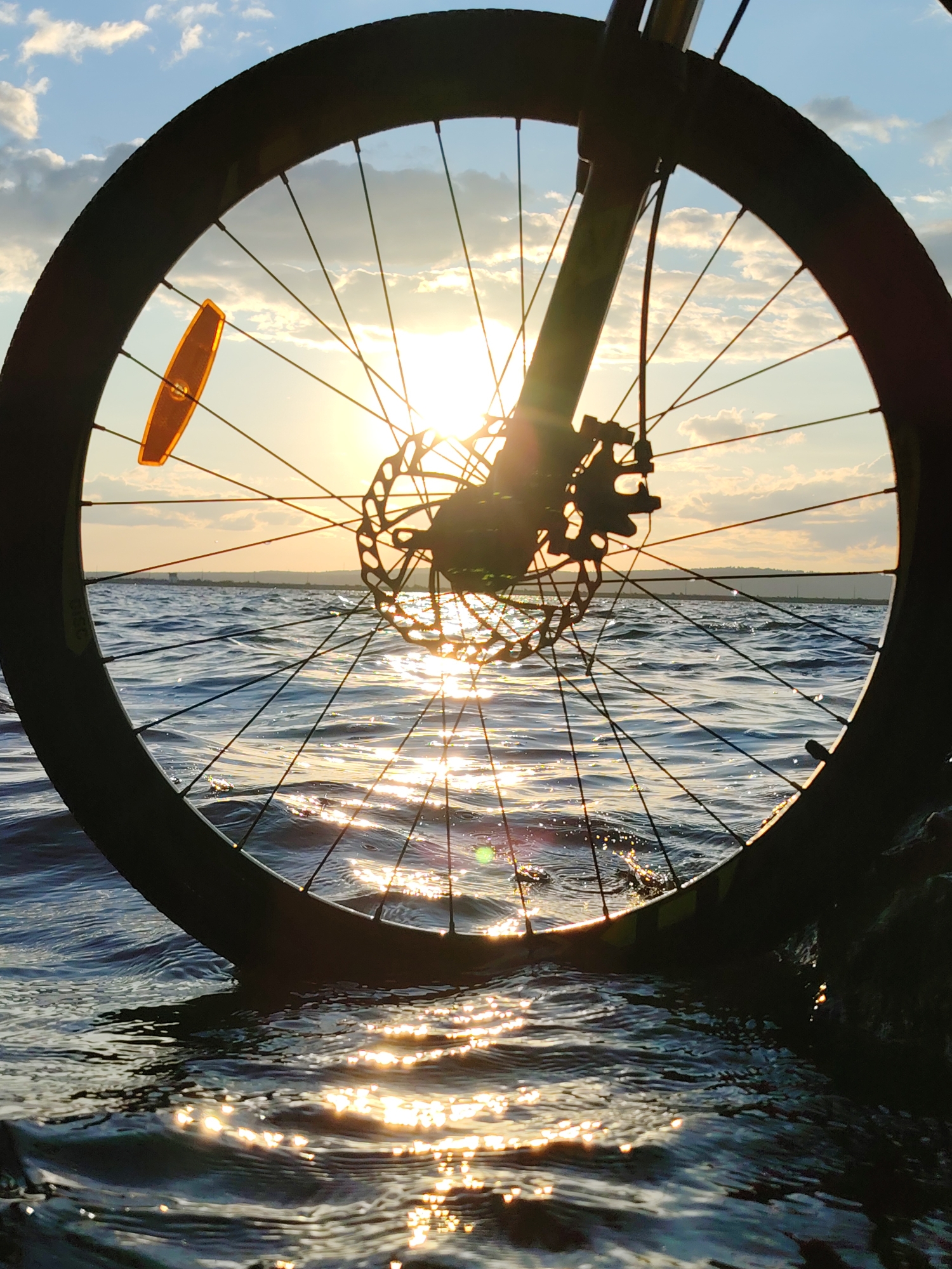 The image captures a bicycle wheel partially submerged in the rippling waters of the Kama River at sunset, near the city of Chaikovsky, Russia. The sun, a bright orb low in the sky, casts a golden shimmer across the water's surface and is visible through the spokes of the wheel. The wheel itself, dark and silhouetted, displays a disc brake mechanism. An orange reflector is attached to one of the spokes. The sky above is a canvas of blue with scattered, wispy clouds. The perspective is close-up, focusing on the wheel and its interaction with the water. The scene suggests a moment of pause or perhaps a dramatic end to a ride. There are no people visible in the image.