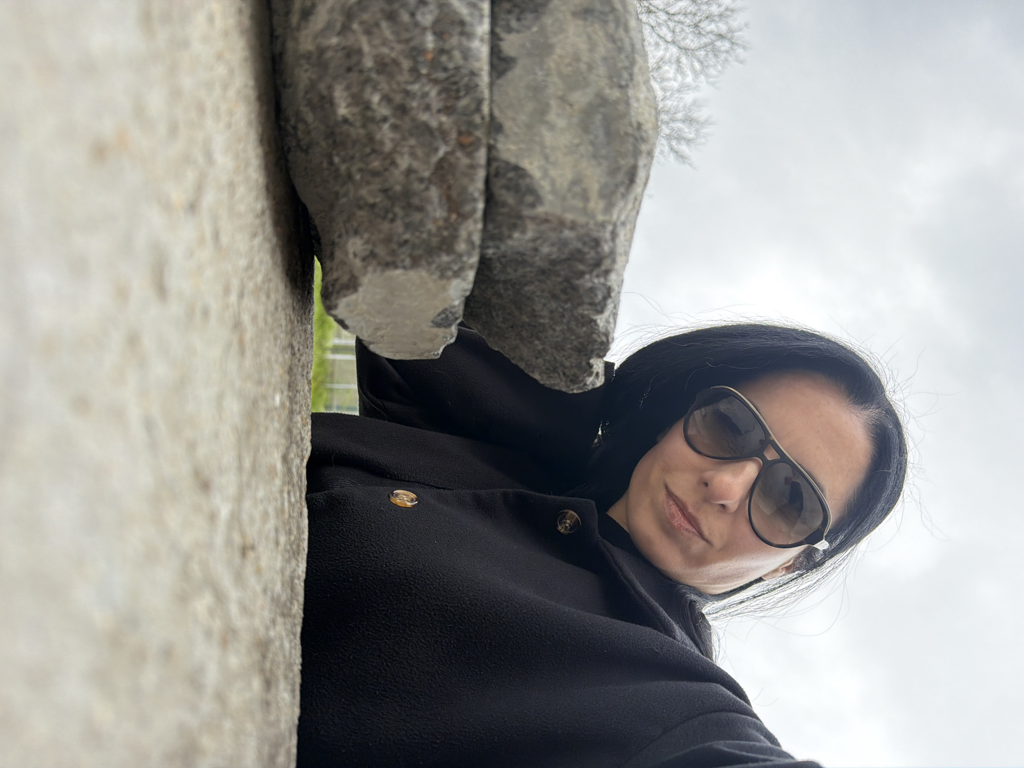 A low-angle shot shows a woman wearing sunglasses and a dark coat standing next to a stone structure. The gray, overcast sky and bare branches of a tree are visible in the background. The stone structure appears to be part of a building or monument, with a rough, aged surface. The location is identified as Pleven, Bulgaria.