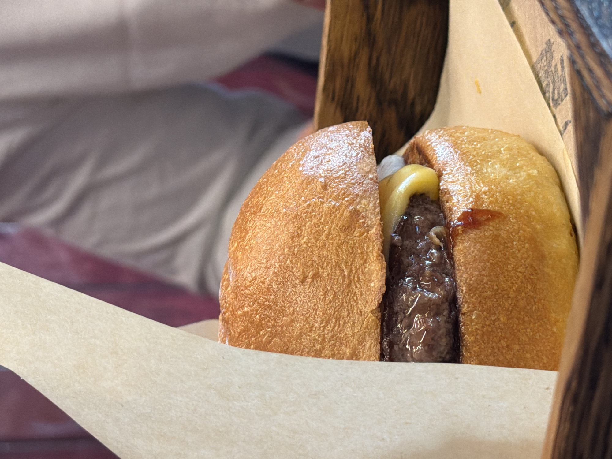 This image captures a close-up of a hamburger, presented in a casual dining environment, specified to be located in Kazan, Russia.
The primary subject is a single, generously sized hamburger, positioned slightly right of center. Its top bun is a golden-brown, toasted brioche-style bun with a somewhat irregular, textured surface. Beneath the top bun, a thick, dark brown beef patty is visible, glistening with what appears to be rendered fat and/or sauce, indicating a juicy texture. A slice of melted, bright yellow cheese drapes over a portion of the patty, with some small, translucent white pieces (likely diced onions) visible underneath it. The bottom bun matches the top in color and texture. A dark reddish-brown sauce or relish is seen dripping down the side of the bottom bun, adding to its visual appeal. The burger is partially encased on its left side by a sleeve of light tan, textured parchment paper, which helps in holding the burger.
The hamburger rests on a larger sheet of similar light tan parchment paper. In the lower right corner of this paper, a faint, dark, indistinct print or logo is barely discernible. This paper, in turn, sits on a dark brown wooden tray or board, exhibiting a visible wood grain.
In the blurred background, to the upper right, the upper torso and arm of a person are visible, wearing a light grey or beige long-sleeved garment. Their body is angled away from the camera. Behind the person, a soft, out-of-focus backdrop consists of a deep reddish-purple area, possibly a wall or upholstered surface, and a lighter, muted grey-beige area adjacent to it. The shallow depth of field keeps the focus sharply on the hamburger. The lighting is soft and even, highlighting the textures and colors of the food.