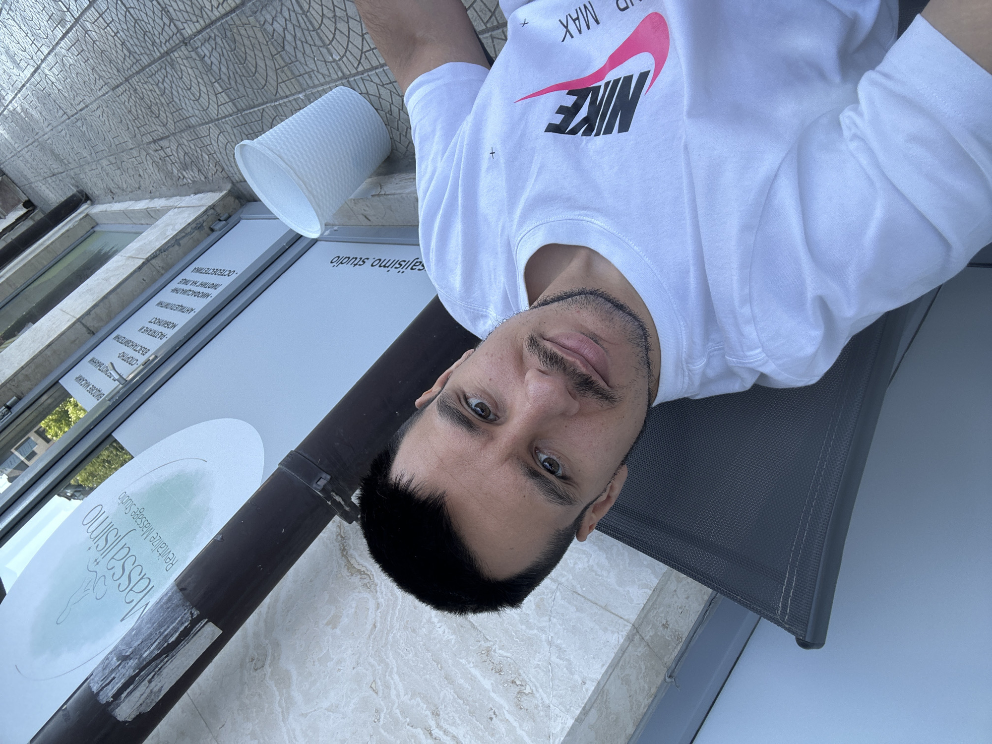 A man is lying on a dark grey lounger, wearing a white T-shirt with a black and pink Nike logo and the words "AIR MAX". He has dark hair, brown eyes, and a mustache. He is looking at the camera. Next to the lounger is a white plastic cup. Behind him is a white sign with the words "Massajisimo Revitalize Massage Studio". The sign also lists different types of massages. The text is written in Bulgarian. Based on the text on the sign "sajisimo, studio" and "Massajisimo Revitalize Massage Studio", and the Bulgarian text, the location is likely in the city of Sofia, Bulgaria. The ground appears to be made of marble tiles. The photograph appears to have been taken outdoors on a sunny day.