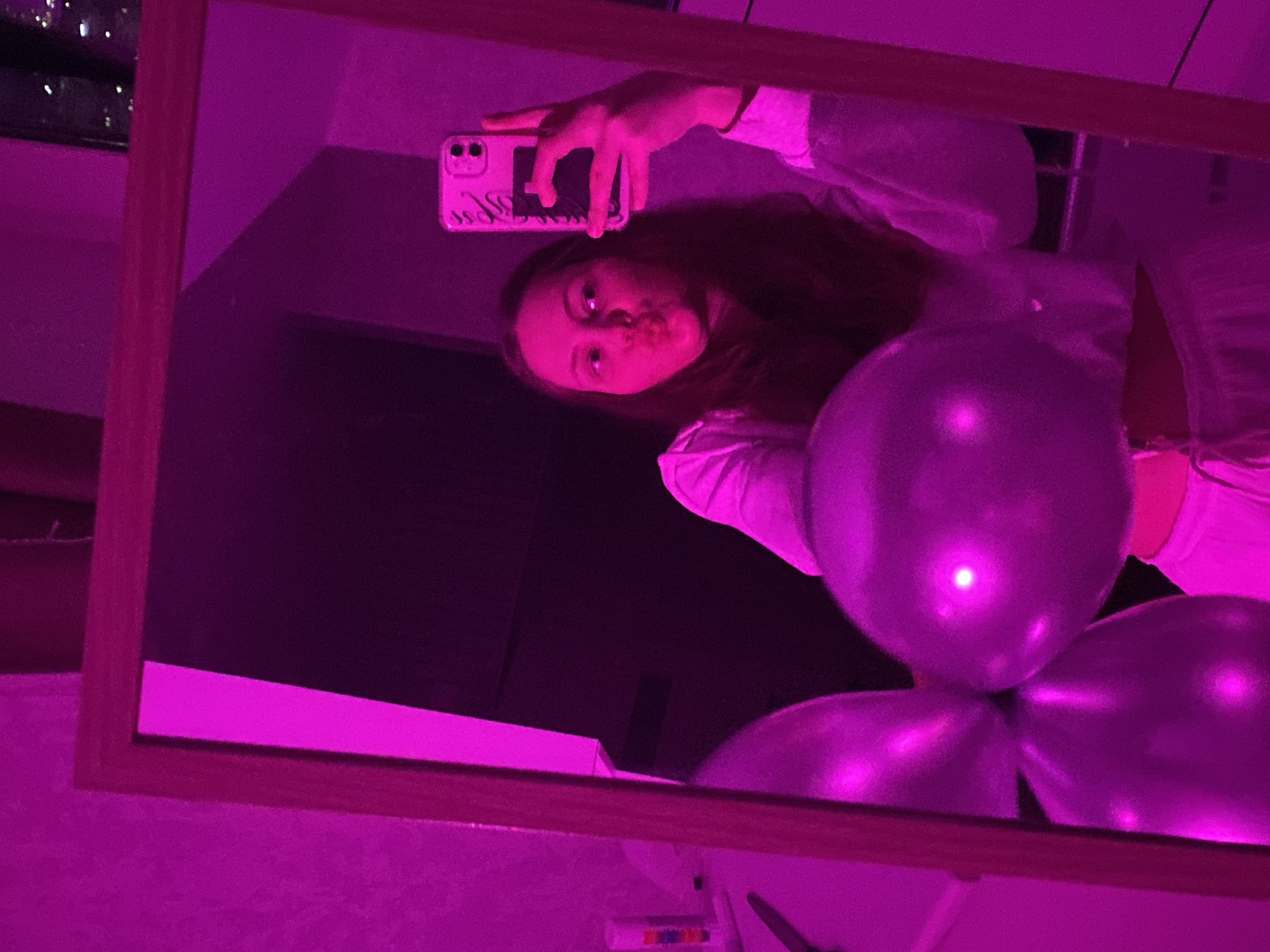 A young woman is taking a selfie in a mirror. The image is bathed in pink light. She is wearing a white top and has her lips pursed as if for a kiss. In her right hand, she holds a smartphone, which is reflecting in the mirror. To her left, partially visible, are several balloons. The background appears to be a room with dark walls and a window showing city lights. The mirror is framed with a wooden border. The location is not identifiable as Izhevsk, Russia.