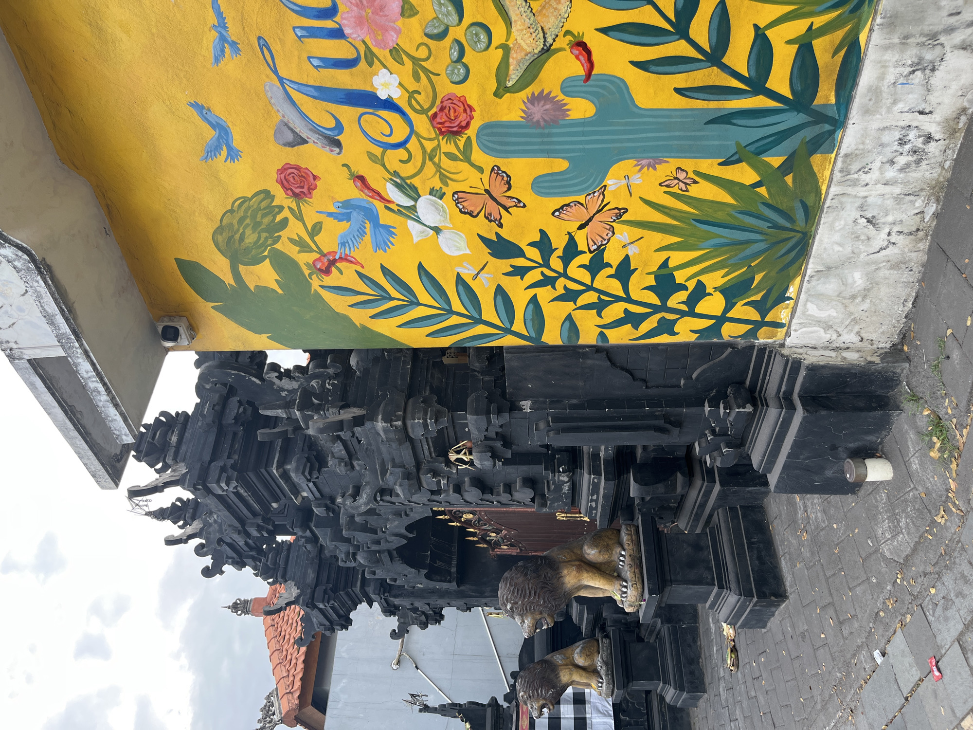 The image shows an ornate black stone temple gate with two lion statues guarding the entrance in Kuta Selatan, Indonesia. The temple gate features intricate carvings and appears to be made of multiple stacked stone blocks. The lion statues are seated on pedestals, facing outward. To the right of the temple gate is a bright yellow wall adorned with a vibrant mural. The mural depicts a collection of colorful elements, including blue birds, red chili peppers, pink and red roses, white flowers, green leaves, and what appears to be a sombrero. There are also images of butterflies, dragonflies, a green cactus, and sliced limes. The ground in front of the temple is paved with dark gray bricks. The sky is overcast.