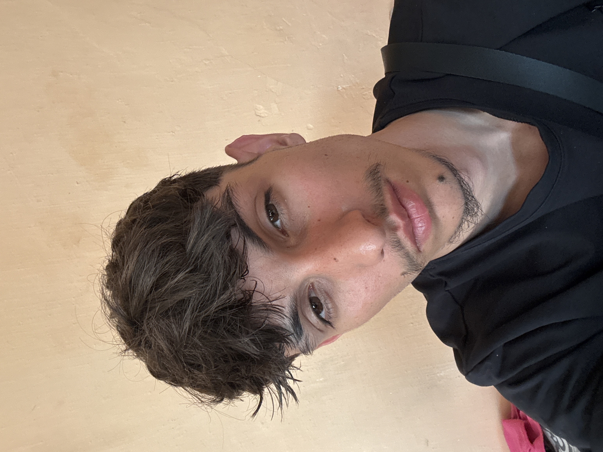 A young man with dark, tousled hair and a light complexion is seen from the chest up, looking directly at the camera. He has a thin mustache and a small beard around his chin, as well as a mole below his lower lip. He is wearing a black t-shirt and a black strap across his shoulder, suggesting he is wearing a bag. The background is a plain, light-colored wall with some texture. The lighting is natural and soft, casting subtle shadows. The image appears to be a selfie taken indoors. The location in Mechkarevo, Bulgaria is not discernible from the image itself.