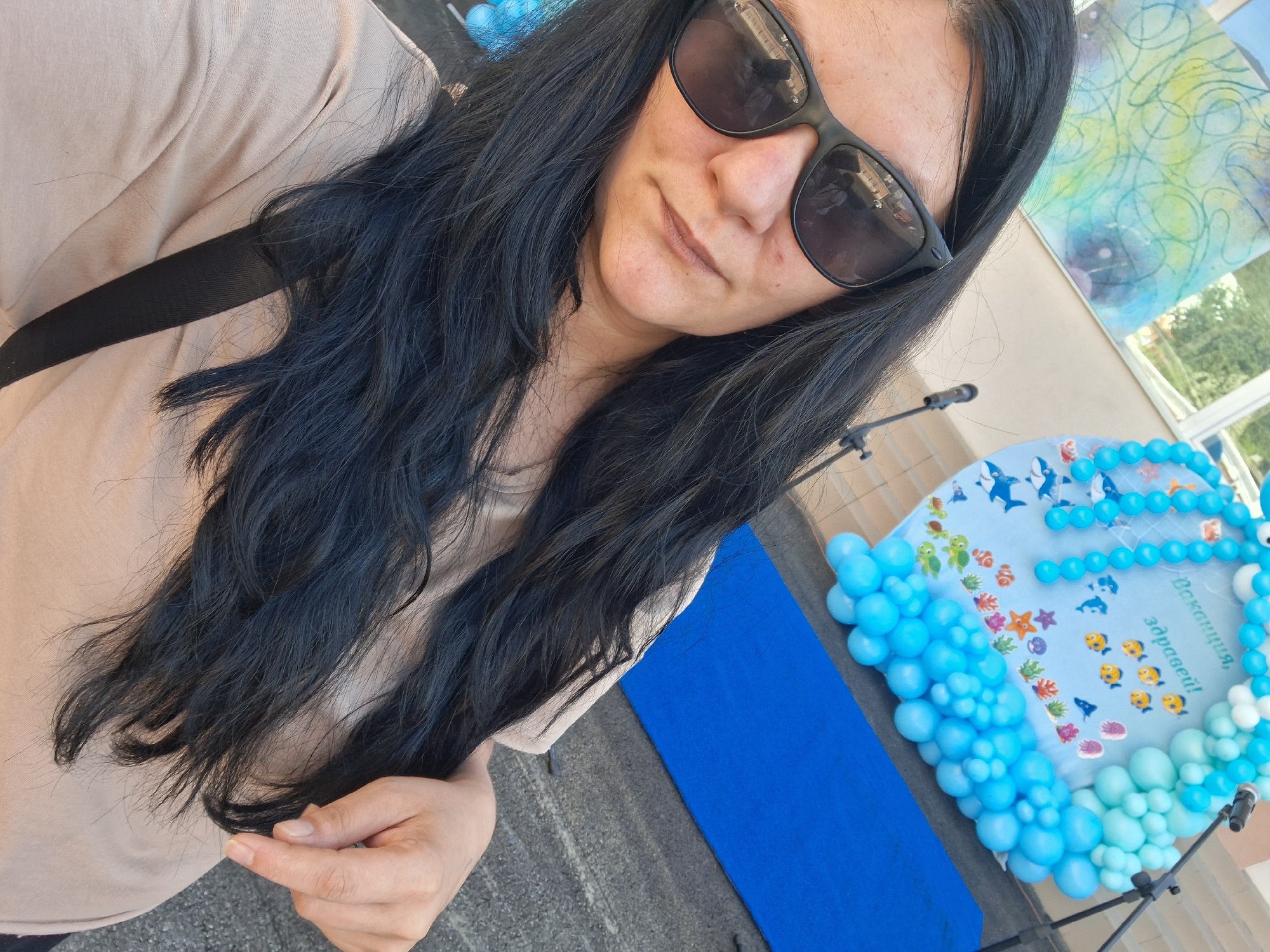In the city of Gotse Delchev, Bulgaria, a woman with long, dark hair and sunglasses is taking a selfie outdoors. She is wearing a light-colored t-shirt and a black strap is visible across her shoulder. In the background, there is a large, blue, circular decoration adorned with balloons and images of marine life, including sharks, dolphins, fish, and turtles. A microphone stand is positioned to the right of the decoration. A bright blue rug lies on the ground in front of the decorated backdrop. The setting appears to be a public space, possibly an outdoor event or celebration.